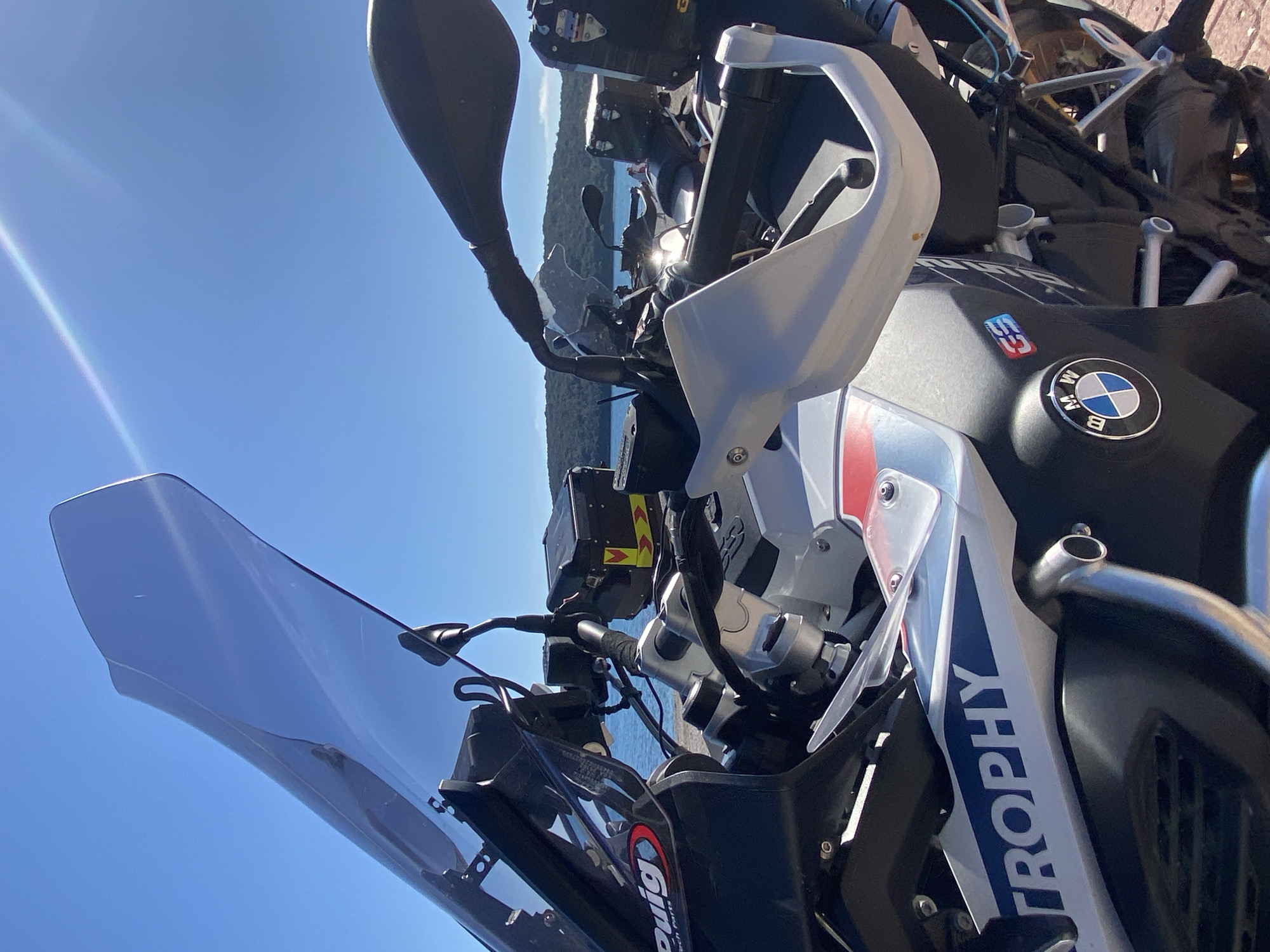 A close-up, slightly rotated shot captures the front of a BMW motorcycle, partially obscuring a view of water and a tree line. The bike features a tall, tinted windscreen, white hand guards, and the "TROPHY" logo in blue lettering on the white fairing. The BMW emblem is visible on the fuel tank, which also displays a "GS" badge. Mounted on the handlebars is an electronic device. In the background, the sea and distant treeline suggest a coastal location. The clear blue sky and bright sunlight indicate a sunny day in Prijevor, Montenegro.