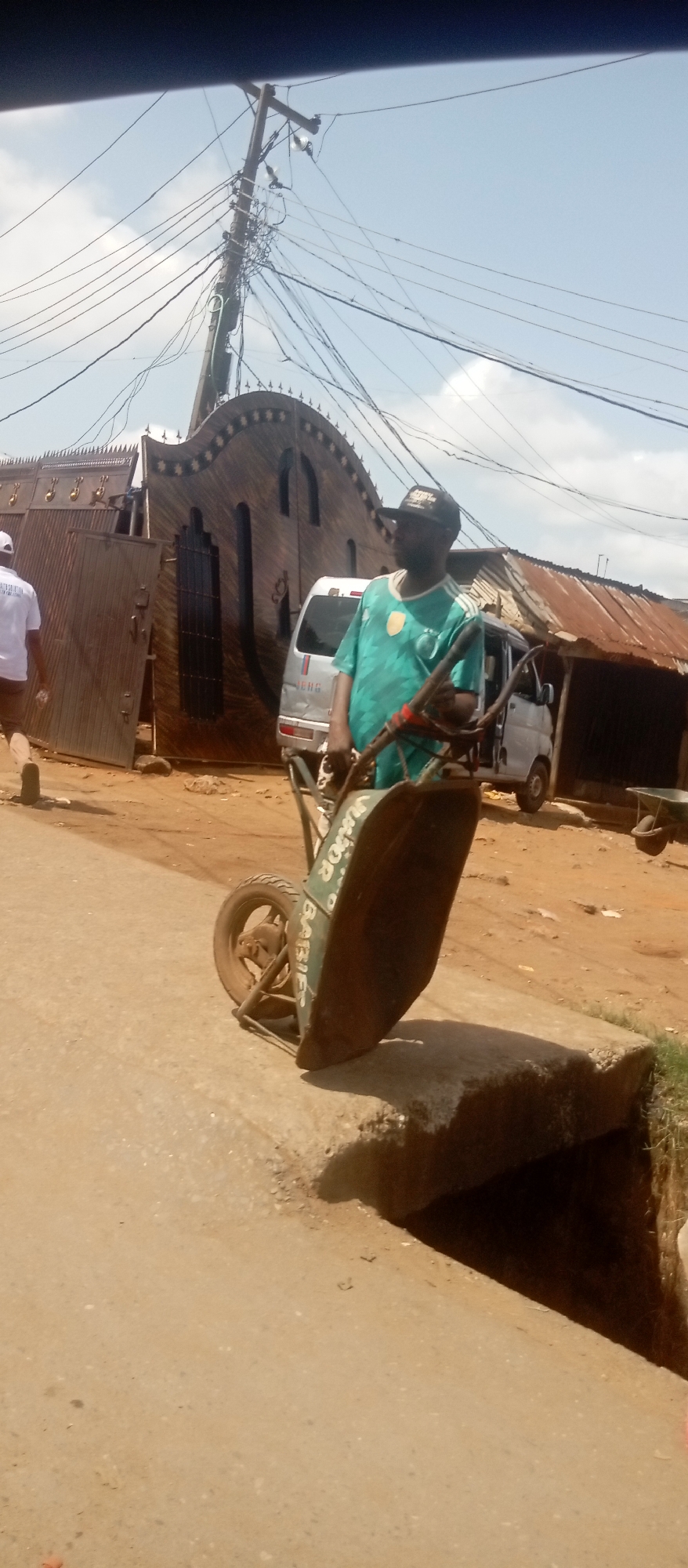 In Onitsha, Nigeria, a man wearing a green shirt and a black cap stands on a concrete ledge with a wheelbarrow. The wheelbarrow is green and labeled "Junior Babies." Behind him, a van is parked in front of a large, ornate wooden gate. An electrical pole with tangled wires is visible above the gate. To the right, there is a building with a corrugated metal roof. A second man, wearing white, walks away from the gate. The road appears to be unpaved. The sky is partially cloudy.