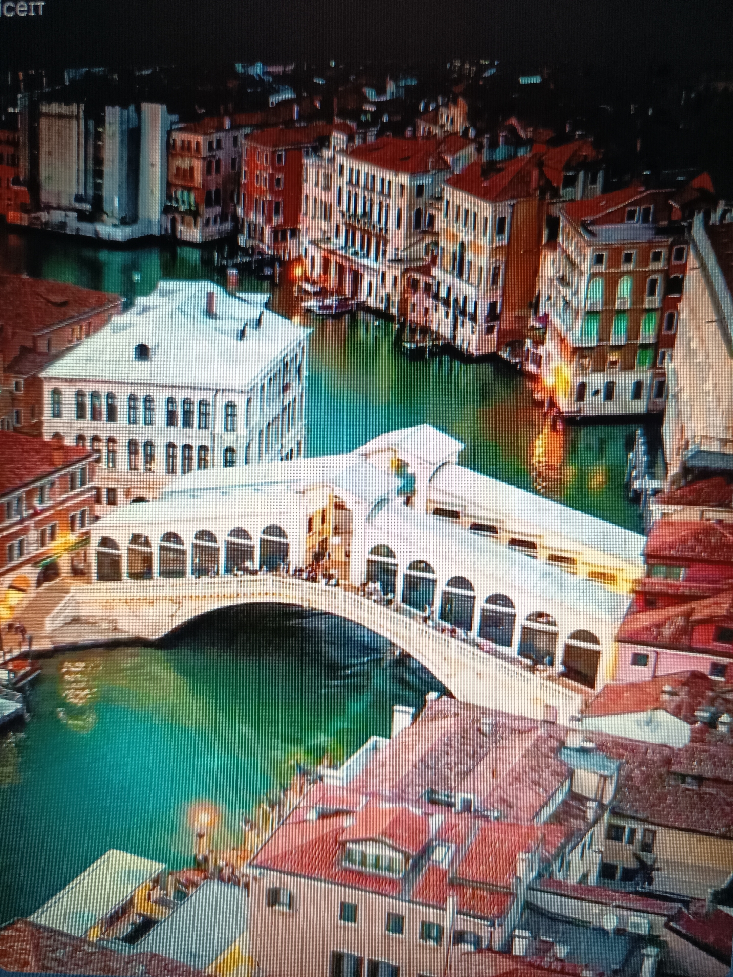 The image captures an aerial view of the Rialto Bridge in Venice, Italy at night. The bridge, a white stone structure with multiple arches, spans a green canal. Shops are visible along the bridge's walkway. Buildings with red-tiled roofs and brightly lit windows line the canals on either side, reflecting in the water. The city is illuminated by artificial lights, creating a warm glow on the architecture and water. A few boats are docked in the canal. The scene is bustling with people visible on the bridge and in the surrounding areas.