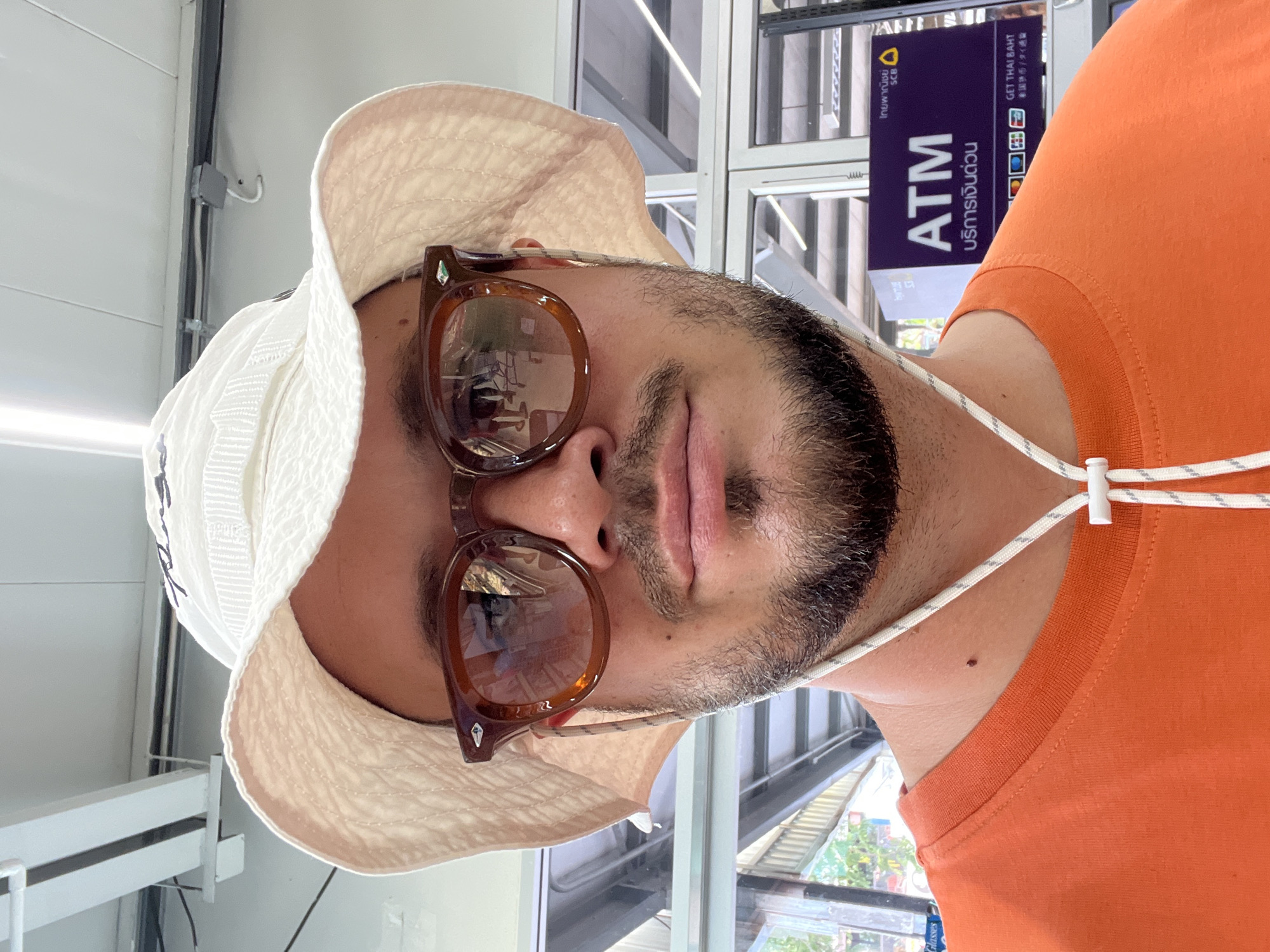 A man wearing a white bucket hat, brown sunglasses, and an orange t-shirt stands inside a building, possibly a bank or commercial space, in an unknown city. In the background, an ATM sign is visible in Thai script, along with a symbol resembling an ATM machine and the letters "ATM".