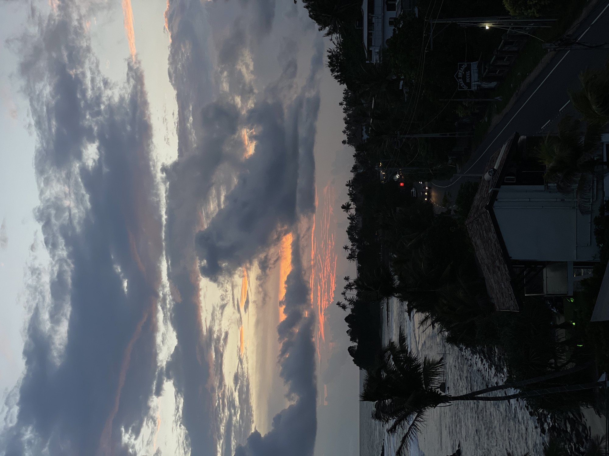 The image shows a view from Goviyapana, Sri Lanka at dusk. The sky is filled with dramatic, dark clouds, illuminated with streaks of orange and pink light from the setting sun. Below the clouds, the landscape includes a coastline with a visible body of water, likely the ocean, and silhouetted trees, including palm trees, along the shore. The right side of the image reveals a road with some streetlights and vehicles; a sign labeled "Ocean Dreams Hotel," and some buildings. The general atmosphere is tranquil, capturing the serene beauty of a tropical sunset in a coastal town.