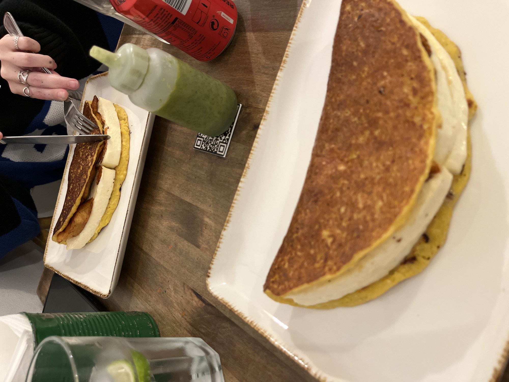 The image depicts a table setting with two plates of food, a green sauce bottle, a red Coca-Cola can, and a glass. A person is cutting into one of the food items with a knife and fork. The food appears to be arepas, corn cakes filled with white cheese. The scene takes place at a table with a wooden surface. The city and location are unknown.