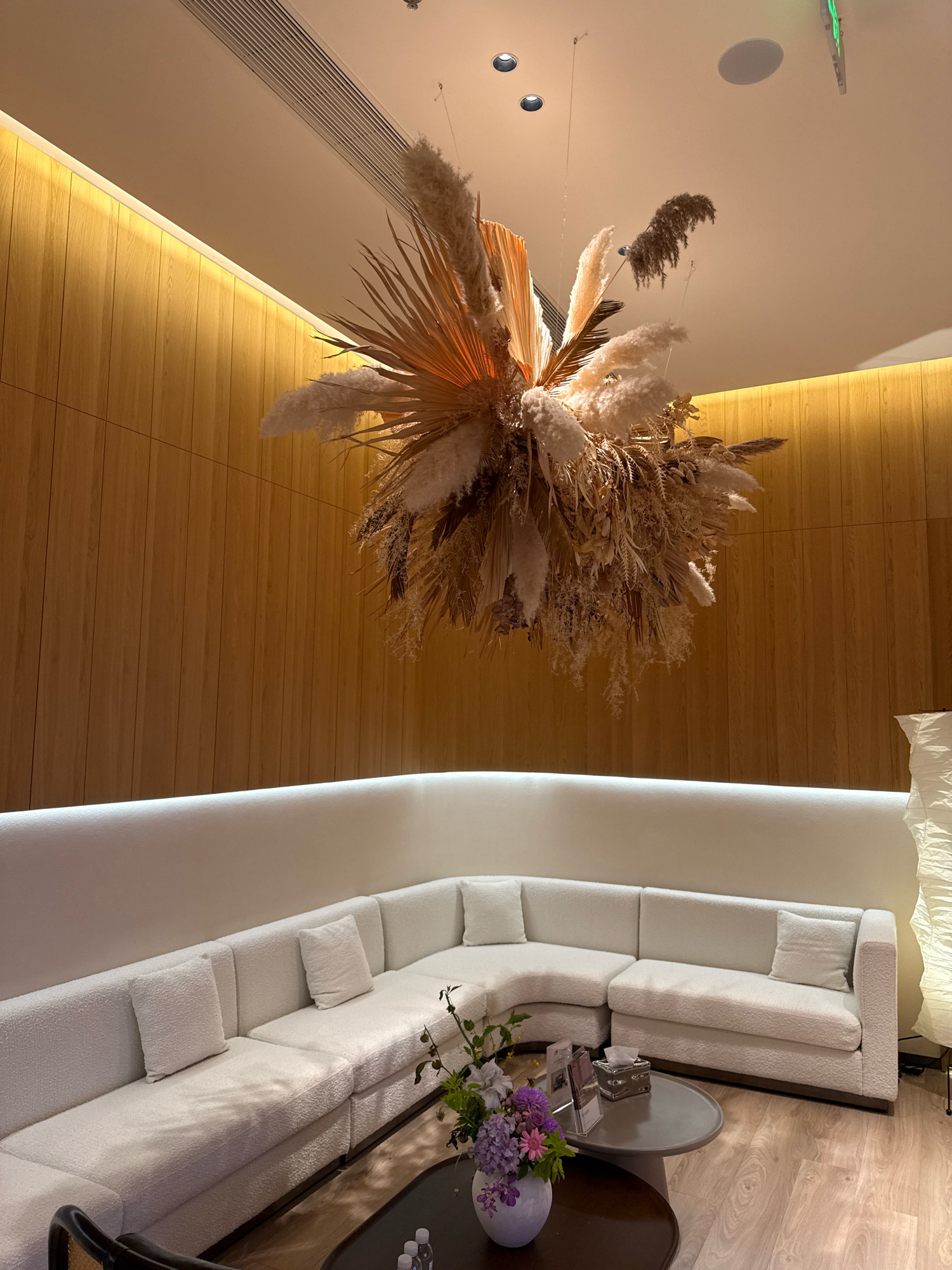 This image depicts a waiting area with a large, L-shaped white sofa featuring textured upholstery and plush cushions. A low, dark coffee table sits in front of the sofa, adorned with a vase of purple and pink flowers, a tissue box, and some papers. To the right of the sofa, a tall, white, textured floor lamp stands. The background features a wall clad in light-colored wood paneling, illuminated by a warm, indirect light strip along the top. Suspended from the ceiling is an elaborate dried floral arrangement. The flooring is light-colored wood laminate. Based on the typical businesses and interior design found in commercial areas of Yiwu, China, this scene is likely located in a boutique hotel lobby, a high-end salon, or a premium retail store's customer lounge within the city. There are no people visible in the image.