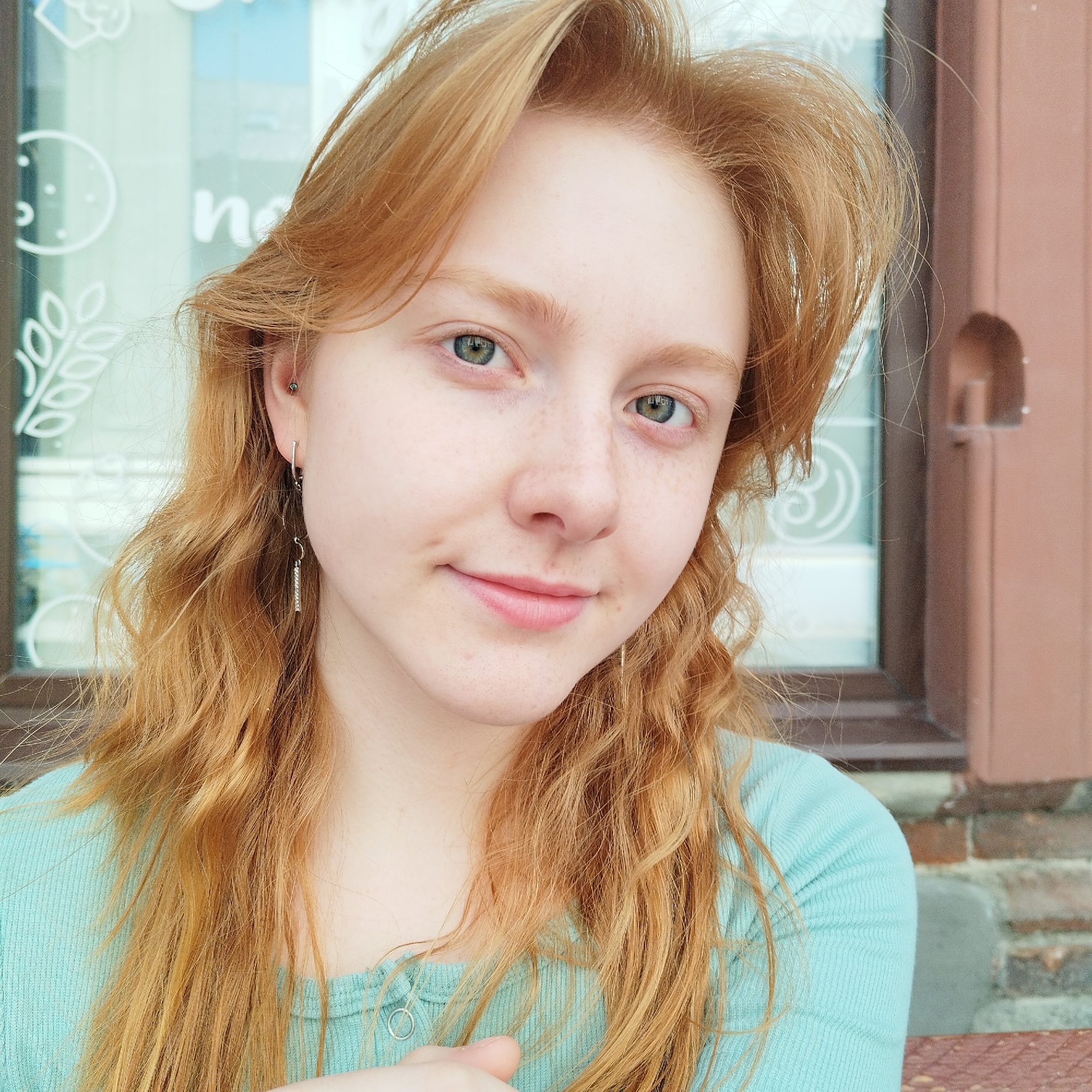 A young person with long, wavy reddish-blonde hair and light green eyes is depicted in a close-up portrait. They have fair skin with freckles across their nose and cheeks and are looking directly at the camera with a gentle, serene smile. The individual is wearing a light blue, ribbed long-sleeved top. On their left ear, they have a small silver hoop earring on the earlobe, a silver dangle earring with a straight bar design, and a small dark stud piercing in the upper helix.
The background behind the person is split. To their left, a large window reveals a bright, slightly blurry interior or outdoor scene. White graphic designs are visible on the window, including a stylized branch of leaves or wheat and a circular shape with dots, resembling a cookie or clock. The letter "n" is clearly visible on the window, part of a larger sign. To the person's right, a reddish-brown brick wall is visible, alongside a darker brown architectural element, likely a part of a building's facade or window frame. The natural lighting suggests it is daytime. The setting appears to be urban, possibly in Yekaterinburg, Russia, near a shop or cafe.
