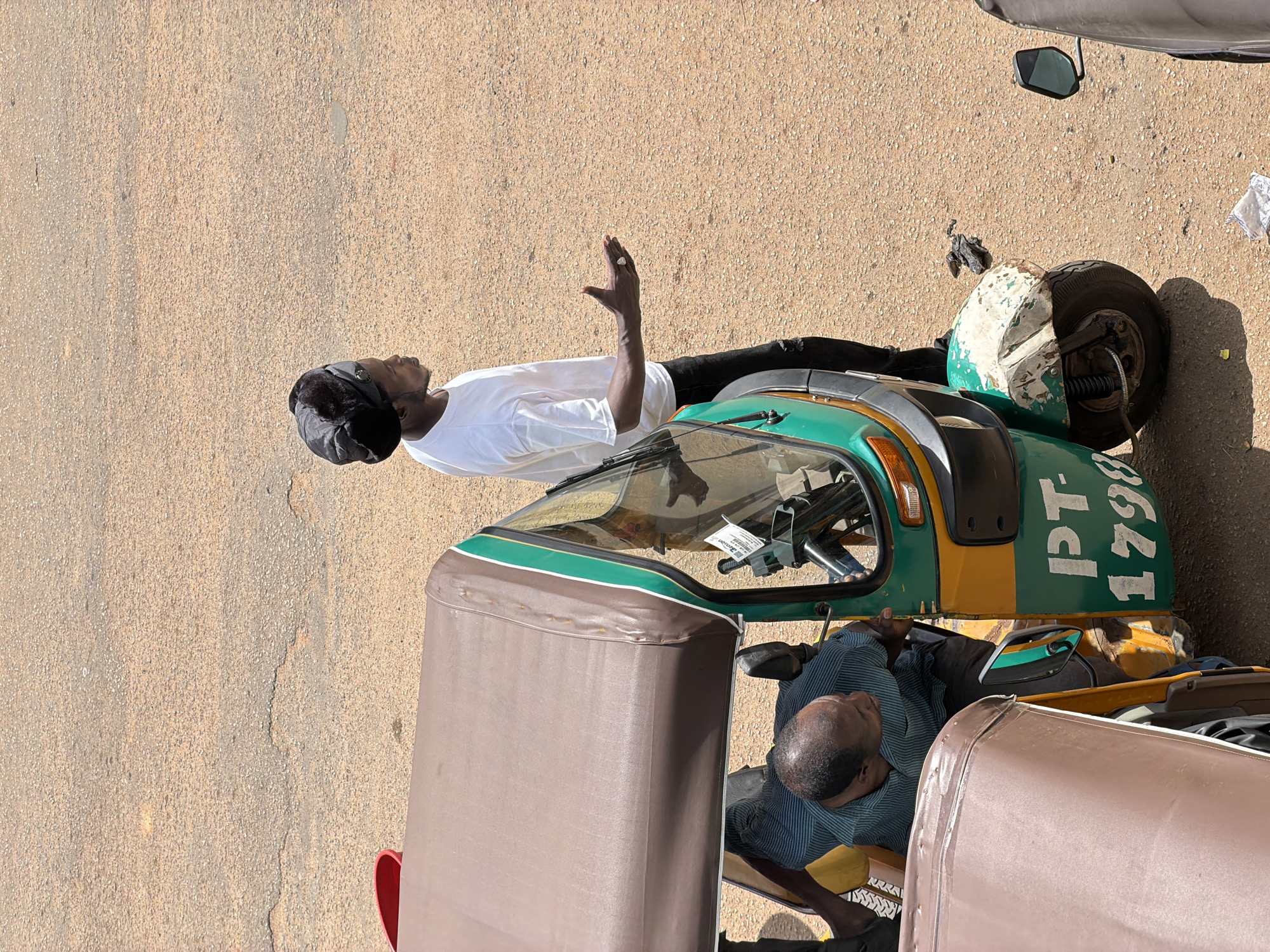 The image shows two men with a three-wheeled vehicle, commonly known as a "keke" in Nigeria. The scene is outdoors on a paved surface, likely a street or road in Bukuru, Nigeria. The ground is a sandy, asphalted texture with some light brown and gray tones, suggesting a dry environment.
One man, wearing a white t-shirt and a black head covering, stands to the left of the keke. He is looking upwards and has his left hand raised, palm facing outwards, in a gesture that could indicate greeting, signaling, or a moment of reflection. He appears to be wearing dark trousers. A ring is visible on his right index finger.
The keke itself is predominantly green and yellow, with some white on its rear fender. The words "PT - 1798" are painted in white on the side of the keke. The vehicle has a canopy and windshield.
The second man is seated inside the keke on the right side. He is wearing a blue and white striped shirt and dark trousers. His head is bowed, and he appears to be resting or sleeping. His face is partially visible, showing a calm expression.
The lighting suggests it is daytime, likely mid-morning or afternoon, given the strong shadows cast by the keke and the man standing. The overall atmosphere seems to be casual and everyday, typical of street scenes in Nigeria. There are no obvious cues about the weather, but the dry ground might suggest it is not currently raining. The image is captured from a slightly elevated angle.