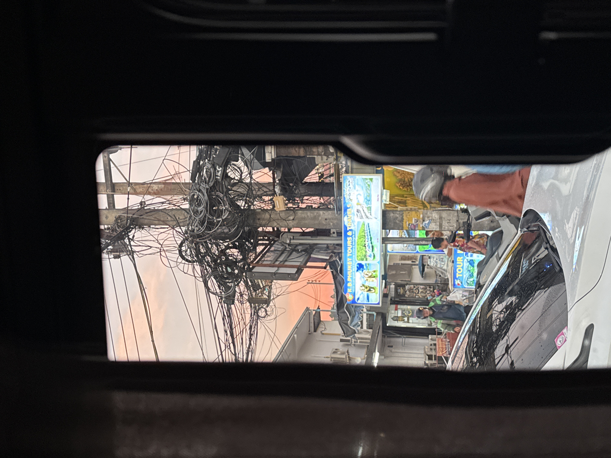 The image, taken from within a vehicle, shows a street scene in Patong, Thailand. Power lines are densely tangled above a street with shops and vendors. People are walking and riding motorcycles. A white car is partially visible on the right side of the frame. Street signs are visible, including one that reads "K.B. PHUKET TOURS & TRE." The sky is light pink.
