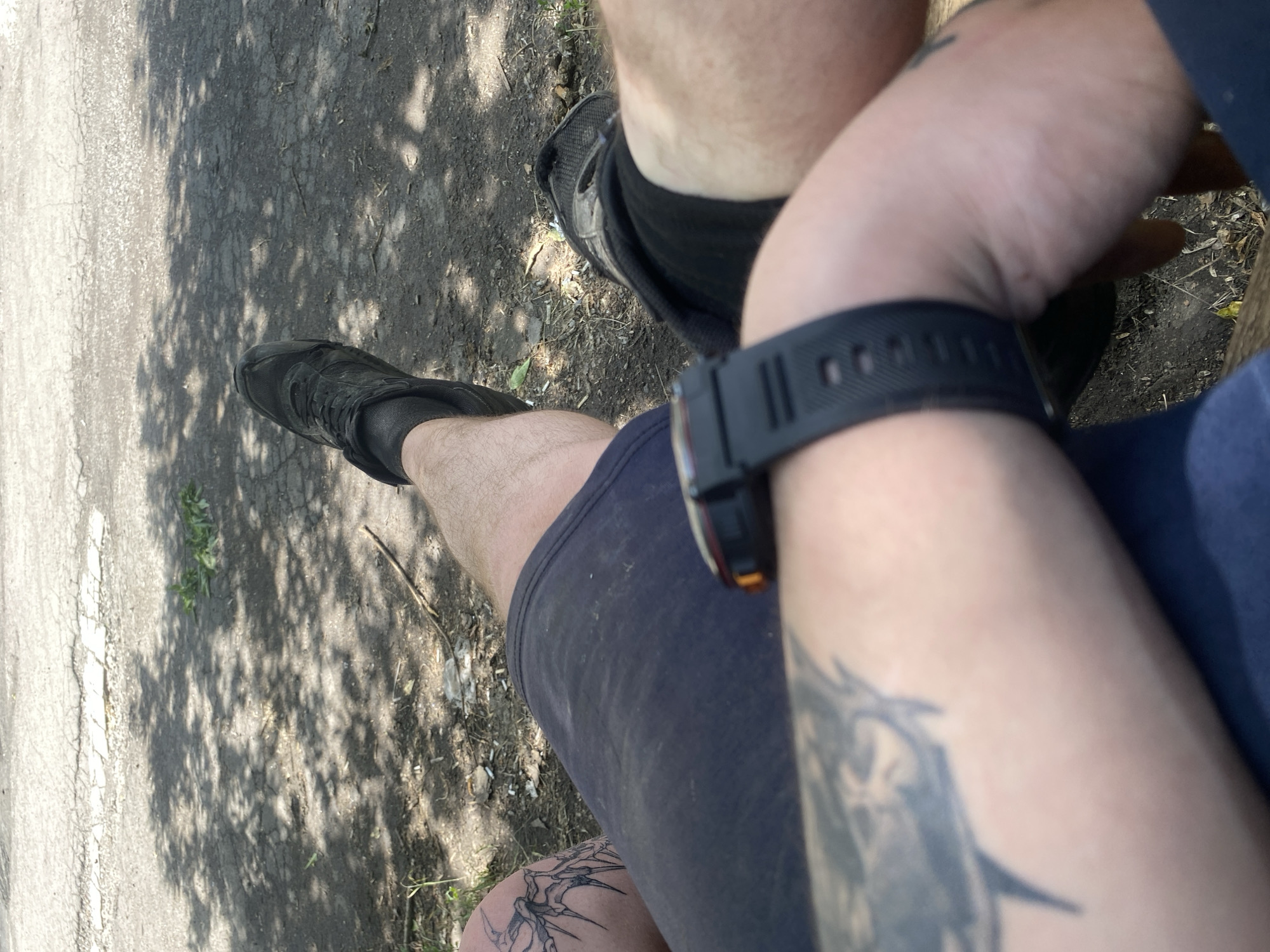 A first-person perspective shot shows a person sitting outdoors, likely on a park bench, with their legs extended forward. The ground is paved with asphalt, cracked and uneven, with shadows cast by unseen trees. The person is wearing dark athletic shorts, black socks, and black athletic shoes. A black smartwatch with a digital display is visible on the person's left wrist. There are tattoos on the person's left thigh and arm, the latter partially visible in the foreground. The environment appears to be a public outdoor space, possibly a park or a recreational area, with some debris such as cigarette butts and twigs scattered on the ground.