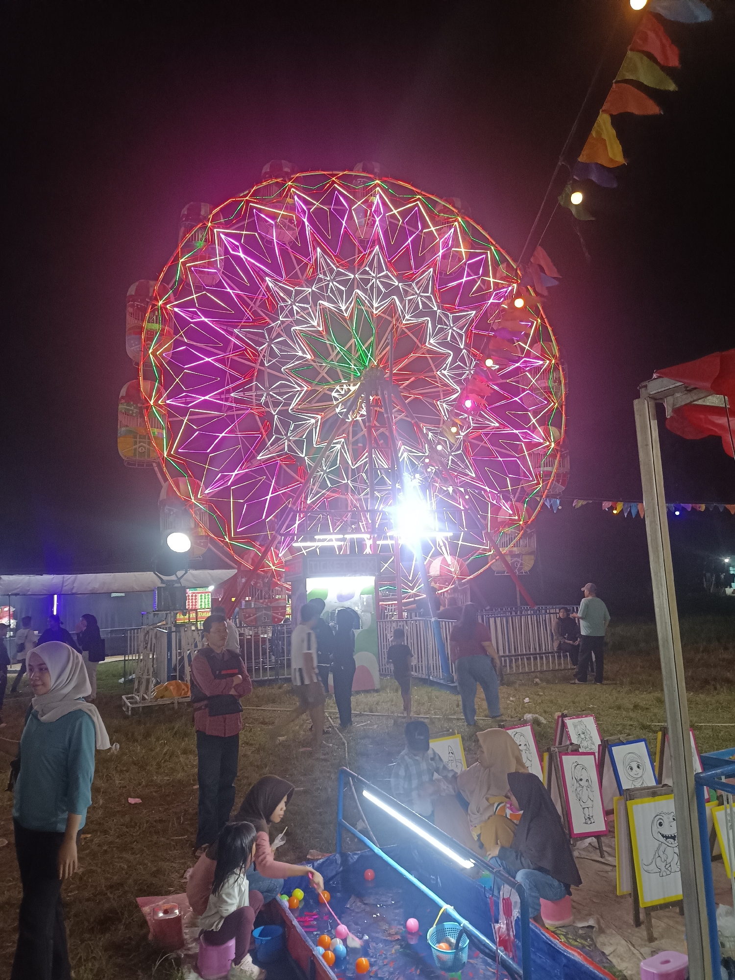 The image depicts a night scene at an outdoor fair or carnival, possibly in Depok, Indonesia. Dominating the background is a brightly lit Ferris wheel adorned with intricate patterns of pink, purple, and white neon lights, creating a festive and vibrant atmosphere. Several gondolas are visible on the wheel.
In the foreground and midground, a variety of people are present, engaged in different activities. Some are standing and observing, while others are seated. To the right, there is a game stall with several children and adults gathered around a water trough filled with colorful balls. Easels displaying drawings of cartoon characters are set up behind the stall, suggesting an art-related activity or prize.
On the left side of the image, a young woman wearing a light blue top and a light grey hijab stands looking to her left. Other individuals are seen scattered throughout the scene, some in casual attire. The ground is covered in grass, indicating an open field setting. Small, colorful flags are strung across poles in the midground, further contributing to the carnival ambiance. The lighting suggests it is nighttime, with the artificial lights of the fair being the primary source of illumination. The overall mood appears to be one of enjoyment and community gathering at a local event. No visible text is clearly legible in the image.