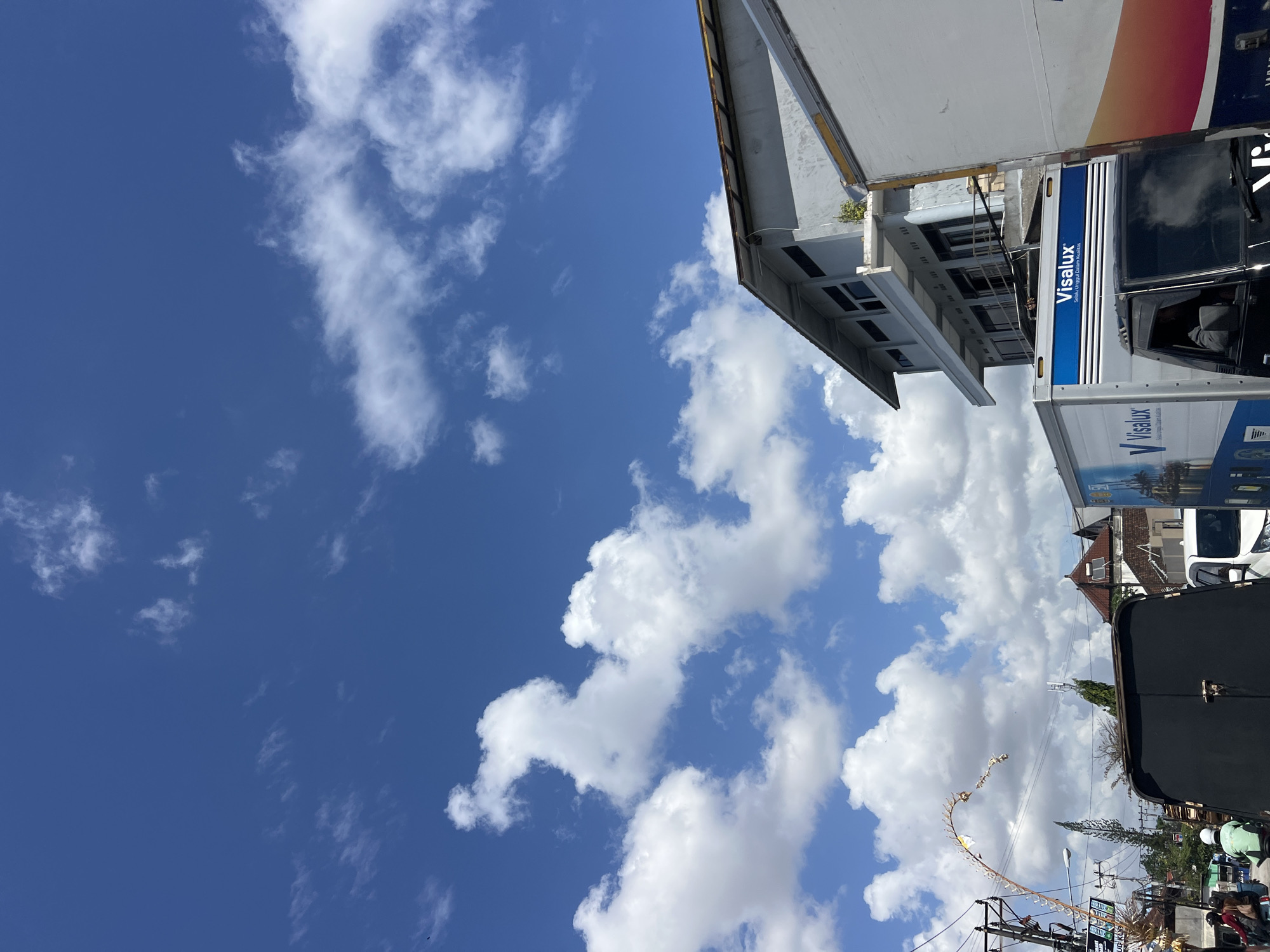 The image captures a street scene in Denpasar, Indonesia, under a bright blue sky dotted with fluffy white clouds. In the foreground, a dark vehicle is partially visible, with a truck labeled "Visalux" to its right. The truck has a blue stripe with the Visalux logo and some text in Indonesian. Further to the right, another truck with a white and orange gradient design is seen.
In the middle ground, a multi-story building with a gray facade and dark windows stands prominently. A traditional Balinese decoration, a long, curved bamboo structure with smaller elements attached, arches over the street from the left side of the image. Power lines and poles are also visible, suggesting an urban environment.
In the background, the roofs of houses with reddish-brown tiles can be seen, along with a tall, slender tree. A few individuals are visible on motorbikes in the lower left portion of the image, one wearing a green jacket and white helmet. The overall impression is a busy street scene on a sunny day in Denpasar.