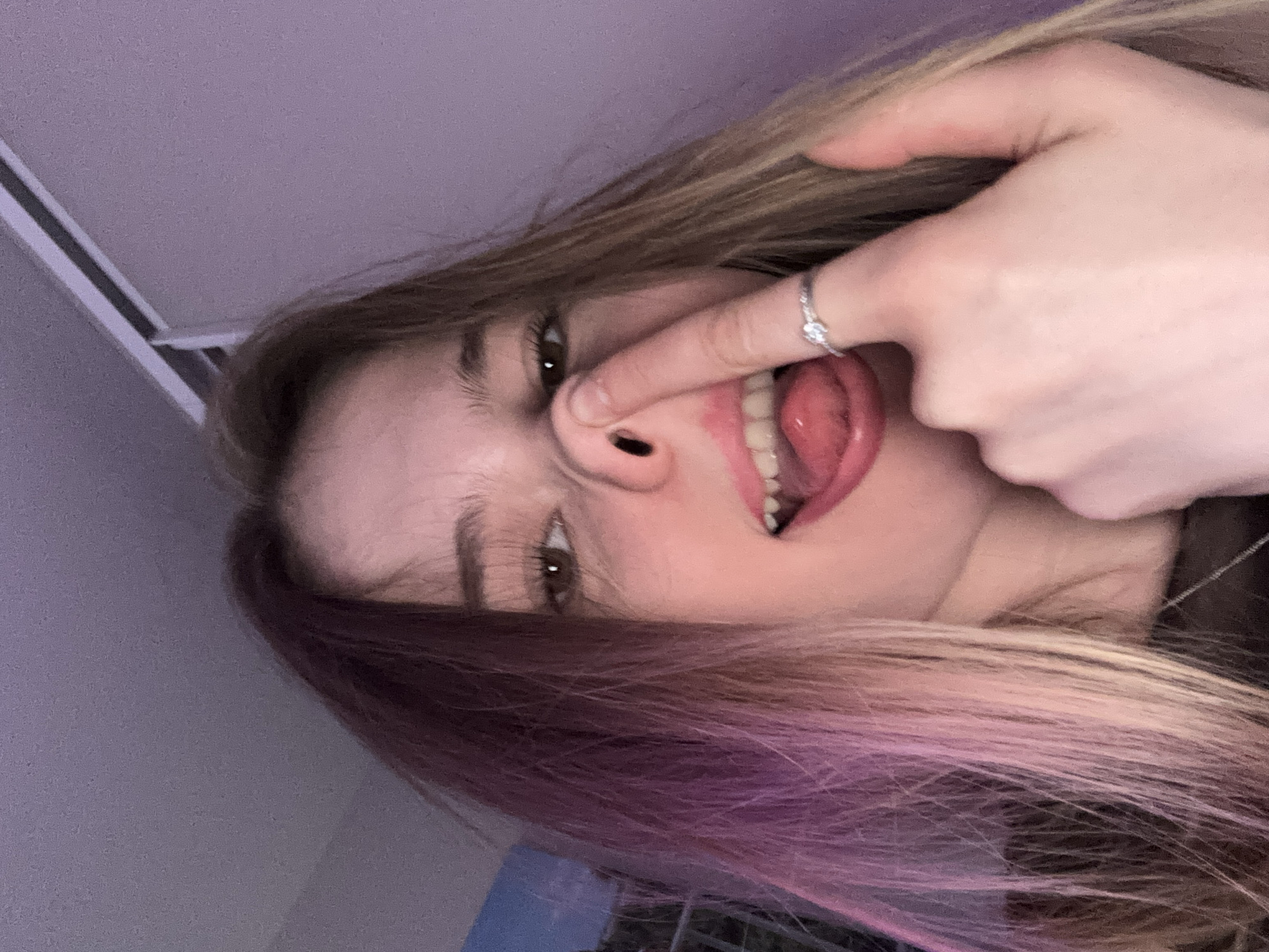 A close-up, rotated portrait captures a young person from a low angle. The subject, who appears to be female, has long hair styled in two distinct colors: the upper section is light brown or blonde, while the lower section, closer to the viewer, transitions into a vibrant purple or pink. She is engaged in a playful act, pressing her left index finger into her right nostril, while her tongue is extended out of her mouth. Her eyes are open and looking towards the camera.
On her left hand, a silver-toned ring with a clear, sparkling gemstone is visible on her ring finger. The lighting is soft, possibly artificial, casting a subtle purple hue on the upper background and her hair. The background is largely out of focus, showing a plain, light-colored ceiling or wall in the upper left, with a light fixture or track running across it. The overall mood conveyed is humorous and lighthearted. No specific location cues are visible within the image to suggest Novoe Devyatkino, Russia, nor is any text present.