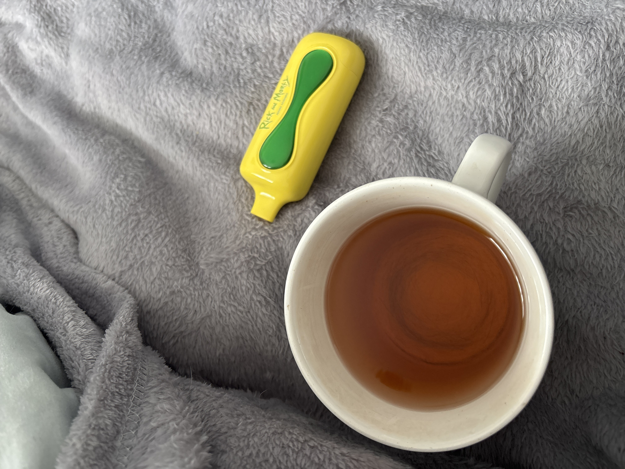 The image shows a top-down view of a gray, plush blanket. On the blanket are two items: a white mug filled with brown liquid (possibly tea) and a yellow highlighter pen with green accents and the "Rick and Morty" logo on it. The location is Unknown, Russia. There are no people present in the image.