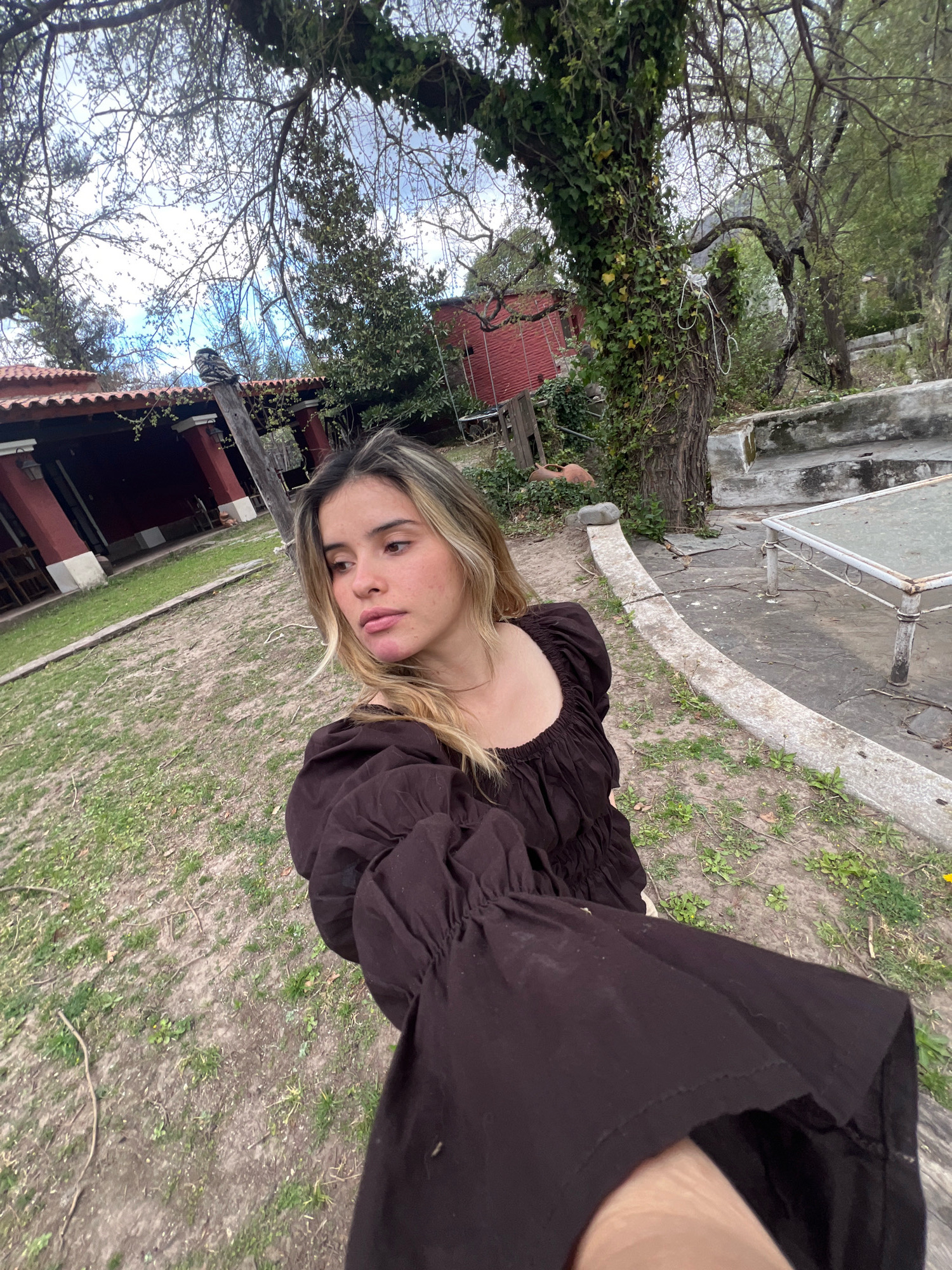 In the city of El Rodeo, Argentina, a young woman with blonde and brown hair, wearing a dark brown ruffled dress, stands in a yard. She is holding her arm up to take a selfie. The yard has patchy grass and dirt, with some dead leaves scattered about. In the background, a red building with a tiled roof is visible, along with several trees, one of which is covered in ivy. A concrete structure with a glass-topped table sits to the right of the woman. The sky is overcast.