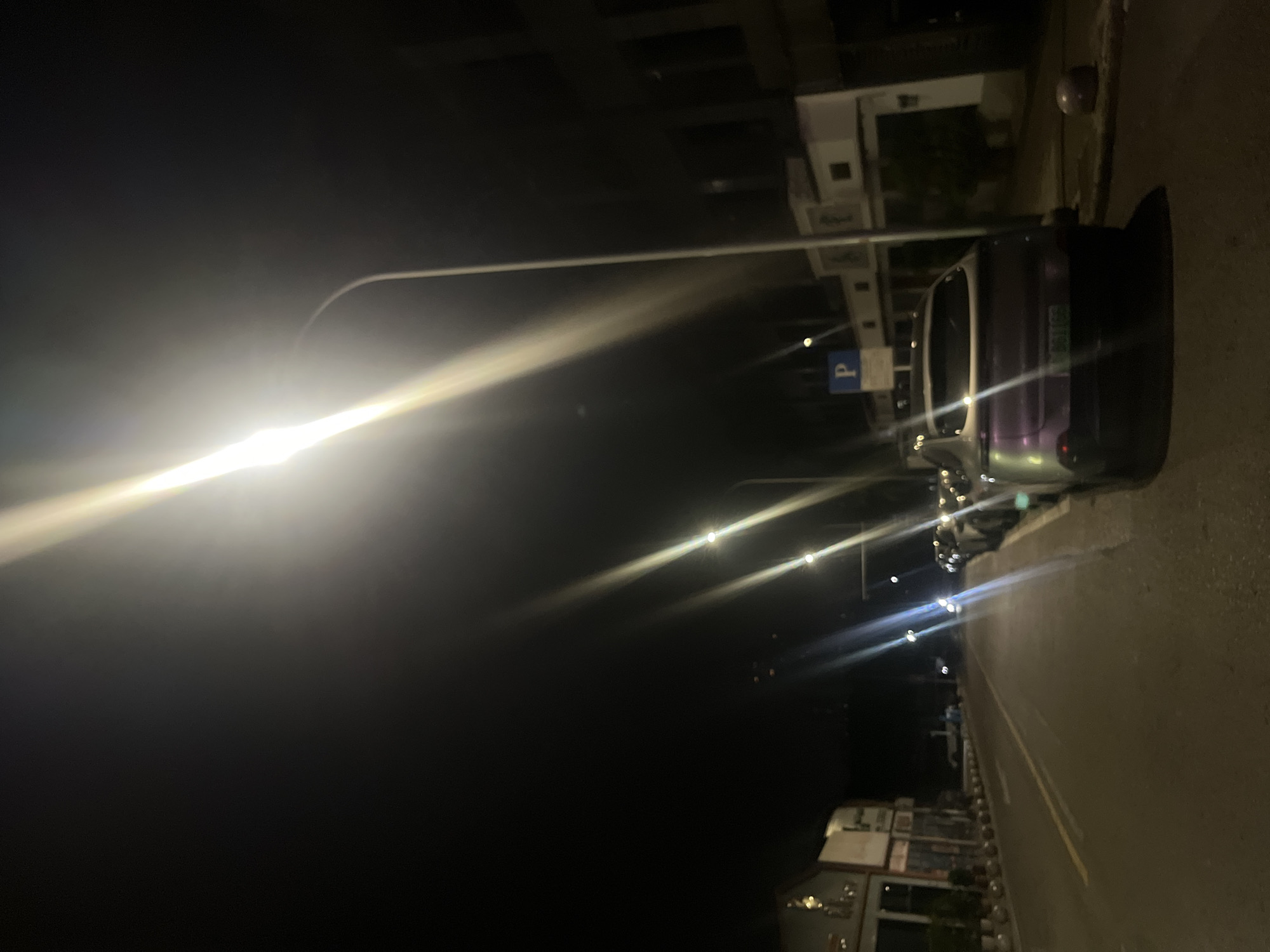 The image captures an urban street scene at night in Jiulongpo District, China. The environment is dark, primarily illuminated by multiple bright streetlights positioned overhead, which cast prominent diagonal lens flares across the upper and central portions of the frame. On the right side of the paved asphalt road, a line of vehicles is parked. The closest vehicle is a light-colored (appearing iridescent purple or silver due to reflection) hatchback or crossover, observed from its rear, with a license plate partially legible as "B611GG." Further down this side, a blue square parking sign with a white 'P' is mounted on a pole, adjacent to what appears to be a low-rise building with a distinctive facade, including an arched structure. On the left side of the road, a continuous row of multi-story buildings, potentially commercial establishments with visible storefronts, stretches into the distance, bordered by a curb and a series of low, spherical bollards. The roadway itself features yellow double solid lines as lane markings. No individuals are present in the visible area of the street.