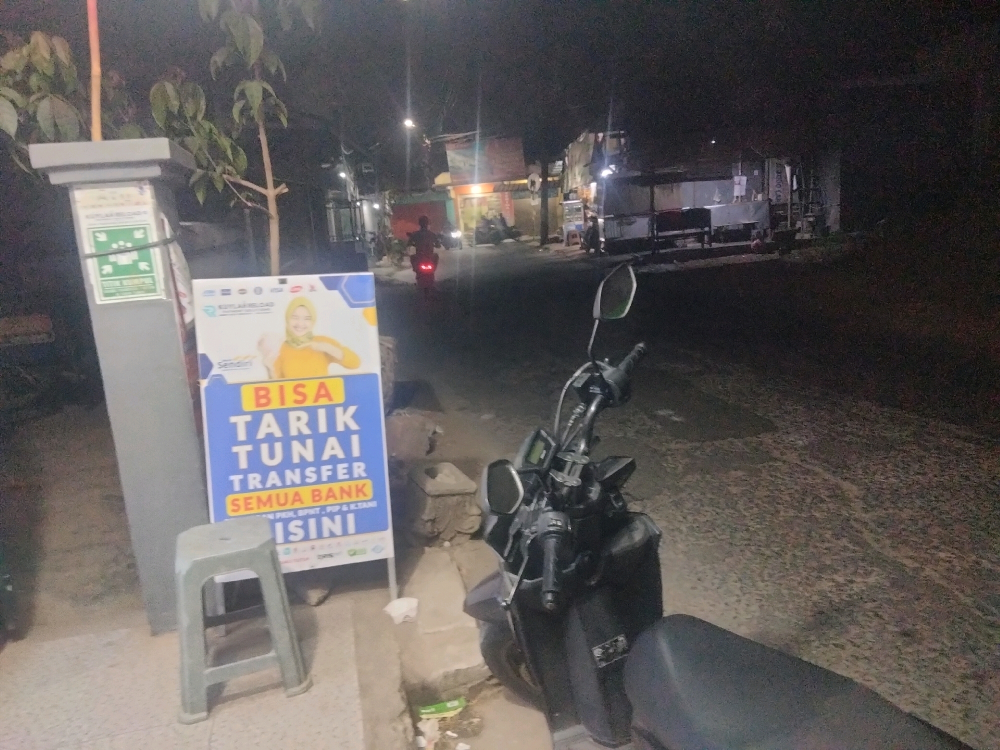 The image captures a street scene at night in Suka Sari, Indonesia. A dark-colored motorcycle is parked prominently in the foreground, its handlebars and front wheel visible. To the left, a gray plastic stool sits in front of a tall, gray pole that supports a sign. This sign has a green background with white text and symbols, and below it, a larger blue and yellow sign advertises financial services in Indonesian, including "BISA TARIK TUNAI TRANSFER SEMUA BANK" (Can Withdraw Cash, Transfer All Banks). The background shows a street with several buildings, some appearing to be shops, illuminated by artificial lights. A motorcycle with a rider is seen in the distance, moving away from the camera. The overall environment suggests a commercial or residential area in a city at night.