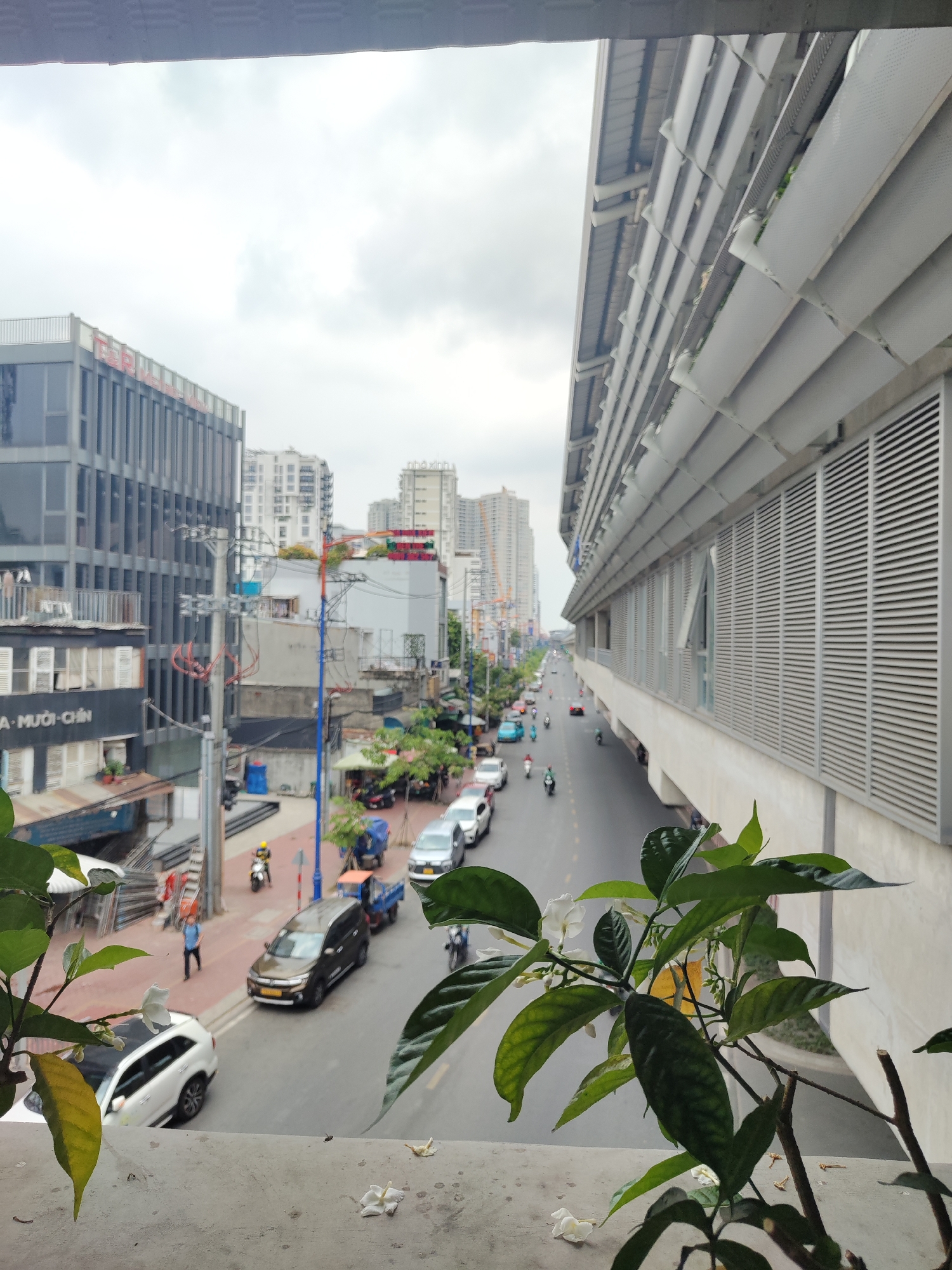 An eye-level shot from what appears to be a balcony overlooks a street in Thủ Đức, Vietnam. The street is lined with buildings of varying heights and architectural styles on the left, with high-rise apartment buildings and structures in the distance. Vehicles, including cars, trucks, and motorbikes, move along the road. Pedestrians can be seen walking along the sidewalks. A plant with white flowers is prominently featured in the foreground, partially obstructing the view. To the right, a large, elevated structure with horizontal metal slats runs parallel to the street. The sky is overcast.