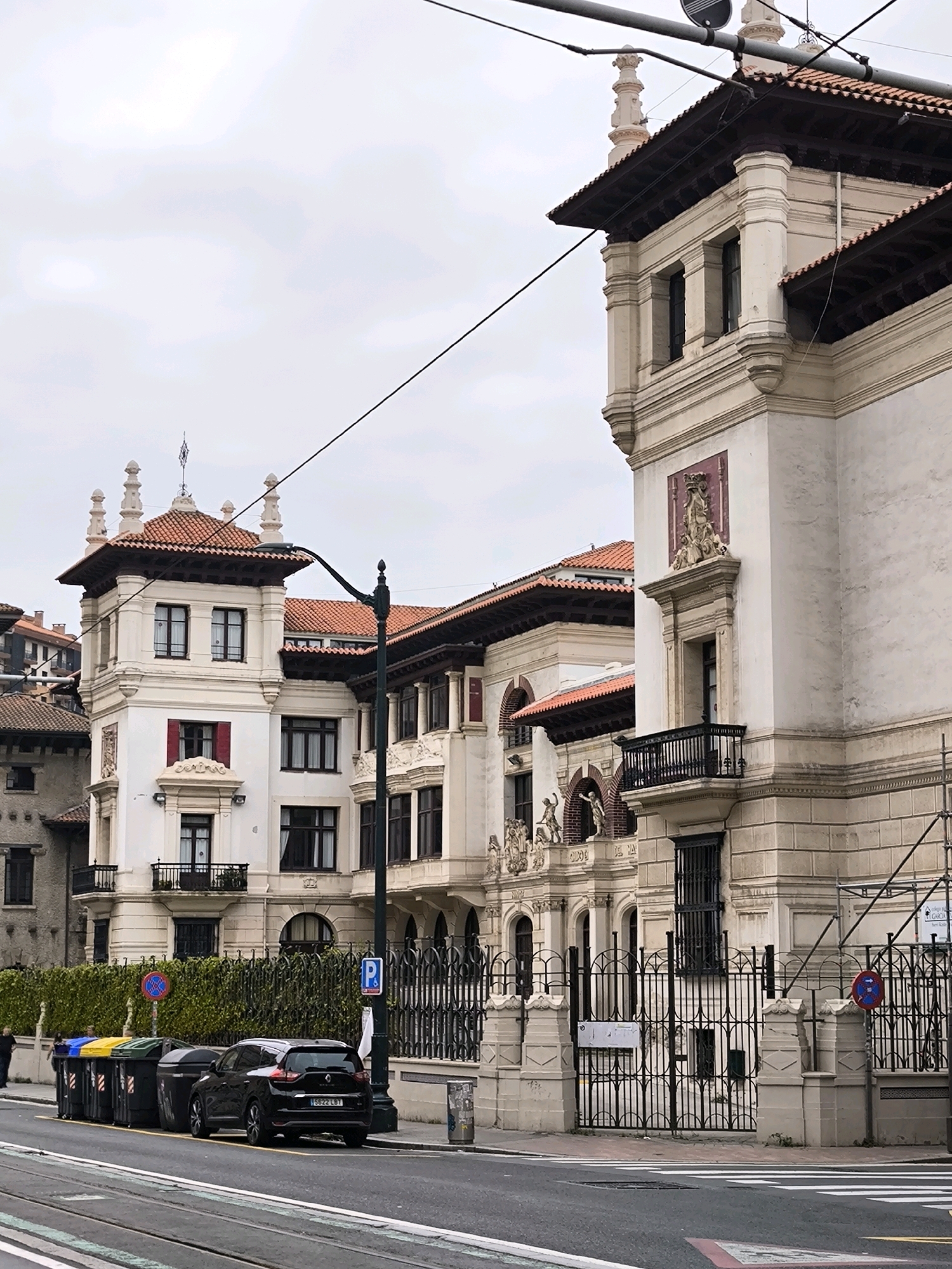 The image shows a street scene in Bilbao, Spain, featuring the striking architecture of the Basque Country Historical Archives building, formerly the City Archives and Public Library. The building, constructed in 1908, exhibits a rich architectural style with ornate facades, red-tiled roofs, and classical ornamentation.
In the foreground, a black SUV is parked along the street next to a row of colorful recycling bins. A pedestrian walkway with a zebra crossing is visible, indicating a busy urban environment. Power lines crisscross the sky above the buildings, a common feature in many cityscapes. The street is paved with asphalt and has visible tram tracks.
The architecture of the buildings is characterized by detailed stone carvings, large windows, and elegant balconies. The building on the right, part of the Archives, displays a decorative relief above a balcony, adding to its historical charm. The building on the left, likely an adjoining structure, shares similar architectural elements, creating a cohesive streetscape.
The overall atmosphere of the scene is one of a well-preserved urban environment with a blend of historical architecture and modern utility. The overcast sky suggests a typical day in Bilbao, known for its temperate maritime climate. The presence of the recycling bins and the parked car grounds the scene in contemporary urban life, contrasting with the timeless elegance of the historical buildings.