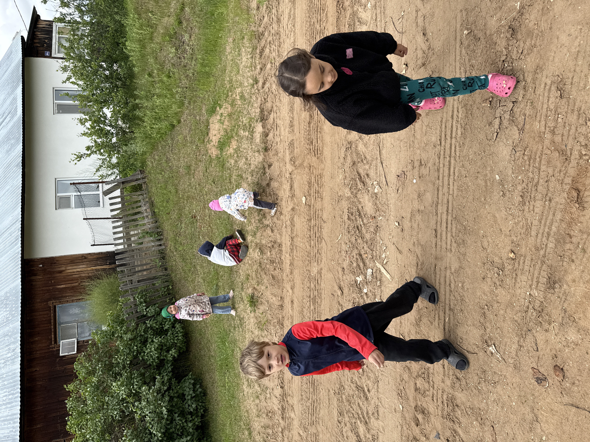 This image captures an outdoor scene in Уральское, Russia, featuring five children on a dirt road and a grassy slope, with two houses in the background under an overcast sky.
In the immediate foreground, a wide, light brown dirt road displays visible tire tracks and scattered small debris. On the left side of the road, a boy, estimated to be 6-8 years old, with light brown hair, walks towards the viewer, slightly angled to his right. He wears a red long-sleeved shirt under a dark blue sleeveless vest, black pants, and dark grey sandals over dark socks. On the right side of the road, a girl, also approximately 6-8 years old, with dark hair styled in two braids, walks away from the viewer, looking downwards. She is dressed in a black fleece jacket, green leggings with a repeating pattern, and bright pink Crocs.
Behind the road, a grassy slope ascends towards the background. On the far left of this slope, a younger child, likely 3-5 years old, stands smiling. This child wears a green knit hat, a light-colored patterned jacket, and light blue jeans. Centered on the slope, another child of similar age sits, facing away from the viewer, wearing a grey and dark blue hooded top and red and black plaid pants. Further right on the slope, a third young child, possibly 2-4 years old, walks away, dressed in a pink knit hat, a light-colored patterned jacket, and dark pants.
The background features two distinct houses. The house on the left is constructed of dark brown wooden planks with a silver-colored corrugated metal roof. It has a window with an open pane and a visible old wooden frame. A large, dense green bush partially obscures this house, and a wooden picket fence runs horizontally across the midground in front of it. The house on the right has white painted walls and a similar silver-colored corrugated metal roof. It features modern white-framed windows, and a small blue sign with "20Б-1" is affixed to its facade. Smaller green bushes are visible in front of this house, along with a section of chain-link fencing.
