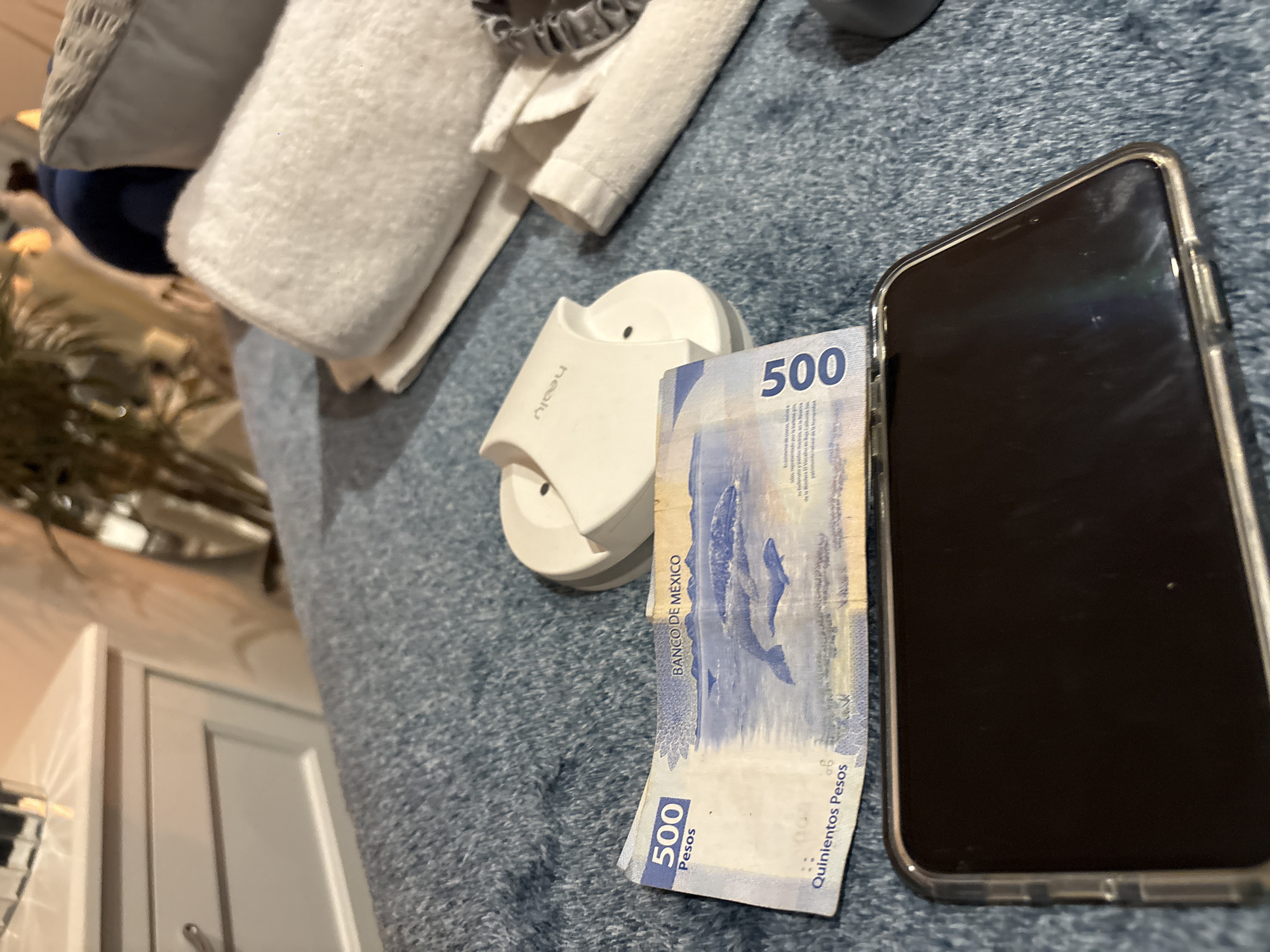 A 500 Mexican Peso bill rests next to a smartphone in a clear case, and a small white electronic device on a blue, textured surface. In the background, there is a stack of white towels and part of a plant. Based on the currency, the location is likely in Mexico. The text on the bill reads "BANCO DE MEXICO" and "Quinientos Pesos" (Five Hundred Pesos). The bill features a whale and text that includes the words "la esfera". It's not possible to determine the specific city or location (e.g., Puebla) from the image alone.