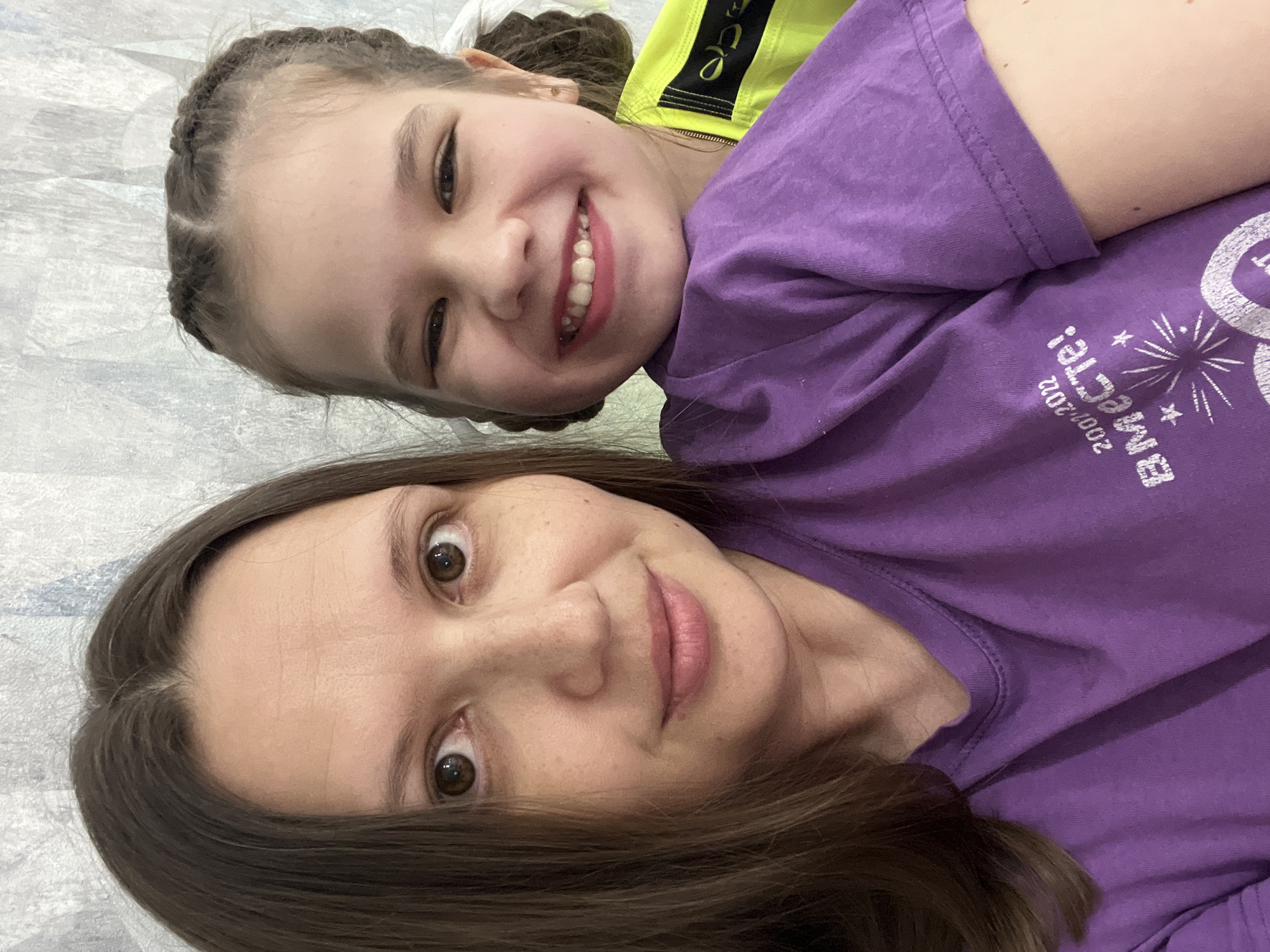 This close-up image features two females, an adult and a child, lying down together and looking towards the camera. The adult is in the foreground, positioned diagonally, with her head resting on a light-colored surface. She has light brown hair, brown eyes, and a calm, pleasant expression, looking directly into the lens. She is wearing a plain purple short-sleeved t-shirt.
Behind and slightly above the adult is the child, who is smiling broadly, revealing her teeth, and looking at the camera with joyful brown eyes. Her light brown hair is styled in cornrows or small braids across her forehead. She wears a bright yellow top under a purple short-sleeved t-shirt. On her purple t-shirt, white Cyrillic text is visible, reading "2007-2022 вместе!" (meaning "2007-2022 together!"), accompanied by a starburst graphic. A small black design or text is also visible on her yellow top.
Both subjects exude happiness and affection, suggesting a close bond, possibly that of a mother and daughter. They appear to be indoors, possibly on a bed or floor, with a light, textured surface serving as the background. The lighting is bright and even. The image captures a candid, joyful moment. While not discernible from the image itself, the location context provided is Chaikovsky, Russia.