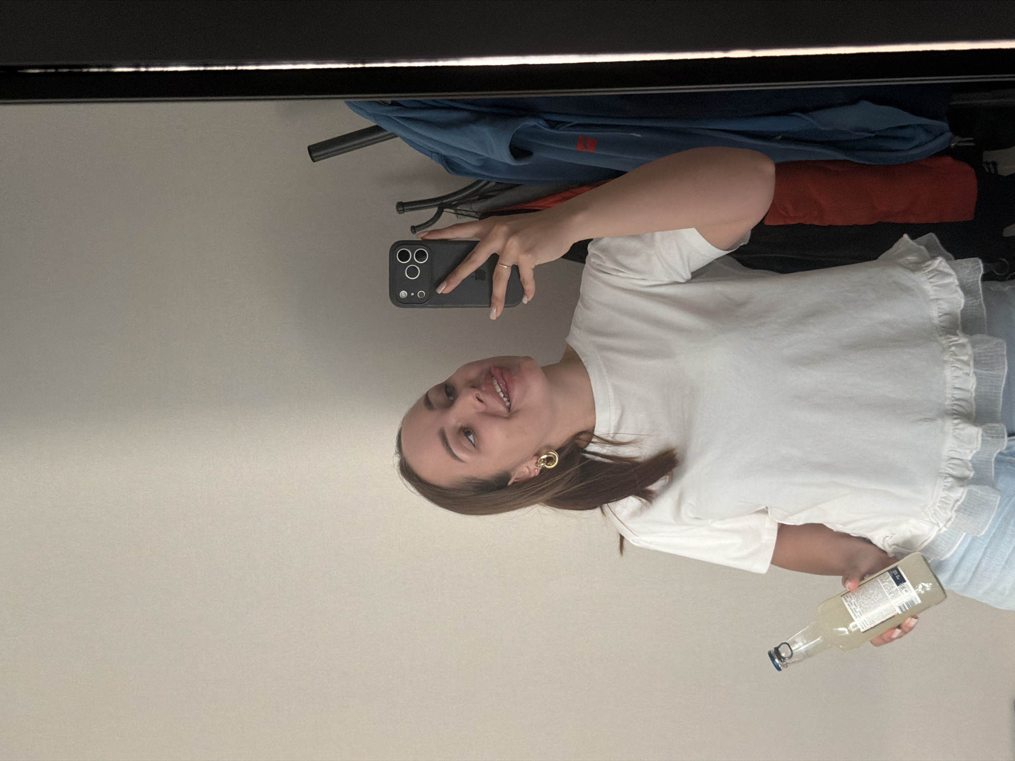 A woman is taking a selfie in a mirror. She is holding a smartphone in her right hand and a bottle in her left hand. She is wearing a white t-shirt, blue jeans and a gold earring in her left ear. A clothes rack with clothing is visible behind her. The location is likely indoors. The location in the city of Kamennoye, Russia cannot be determined based on the visual data.