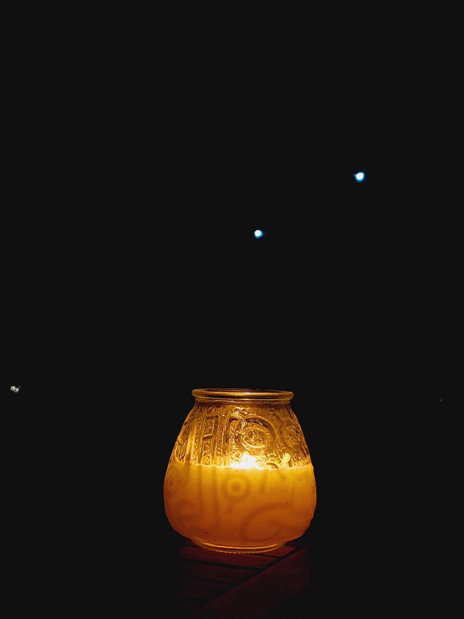 A lit candle in a decorative glass jar sits on a dark surface. The background is entirely black, with three small, bright blue lights scattered in the upper portion of the image. The candle emits a warm, yellow glow that illuminates the textured glass of the jar, revealing intricate patterns. The bottom of the jar rests on what appears to be a dark wooden surface. The overall impression is one of quiet, ambient lighting against a dark backdrop. There are no people or animals present. The location is not discernible from the image itself, and there is no information to link it to Sozopol, Bulgaria.