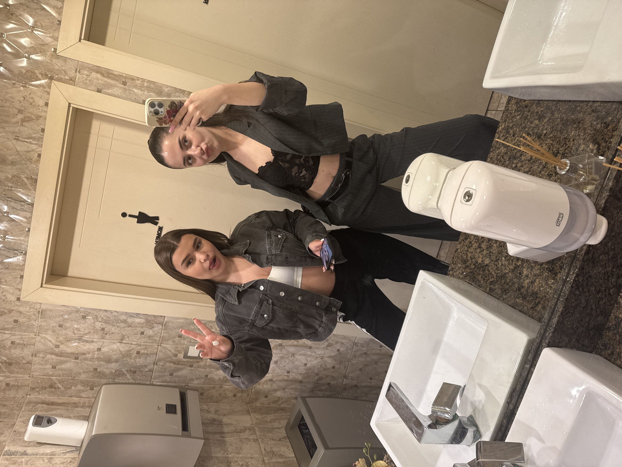 Two young women are captured in a mirror selfie within a public restroom, located in Targovishte, Bulgaria. The image is oriented vertically, showing the women posing in front of sinks and tiled walls.
The woman on the left, who is actively taking the photo with a phone in a distinctive teddy bear and floral case, has dark hair pulled back. She wears a dark gray pinstriped blazer over a black lace bralette, paired with matching trousers. She looks directly at the camera with a slightly pouty expression.
The second woman, positioned to the right, has long dark hair and is dressed in a dark gray denim jacket, a white crop top, and black pants with a white stripe down the side. She holds a phone in her left hand and gestures a peace sign with her right, displaying a playful expression with her tongue slightly out.
The background reveals a well-maintained public restroom environment. Multiple white rectangular sinks with chrome faucets are set into a dark speckled granite countertop. The walls are covered with light brown tiles featuring a raised geometric pattern. Visible fixtures include two white wall-mounted soap dispensers, one clearly labeled "TORK," and a white paper towel dispenser. A small dark gray trash can and a glass diffuser with reeds are also present. In the upper background, two light-colored wooden doors are visible, one bearing a partial "WOMEN" restroom sign (showing "MOMEN" and an inverted stick figure icon). The lighting is artificial, suggesting an indoor setting, likely in the evening or at night.