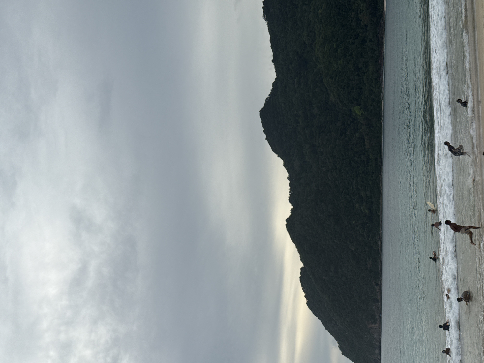 The image depicts a beach scene with a lush green mountainside forming a dramatic backdrop. The sky is overcast with soft, diffused light, suggesting it's either early morning or late afternoon. The ocean is a calm turquoise, with gentle waves breaking on the sandy shore. Several people are scattered along the water's edge, some wading in the shallows, while others are standing or sitting on the sand. A few individuals appear to be engaging in water activities, possibly surfing, as surfboards are visible. The overall mood is tranquil and relaxed, with the natural beauty of the location taking center stage. The scene is set in Meunasahbale, Indonesia. There is no visible text in the image.