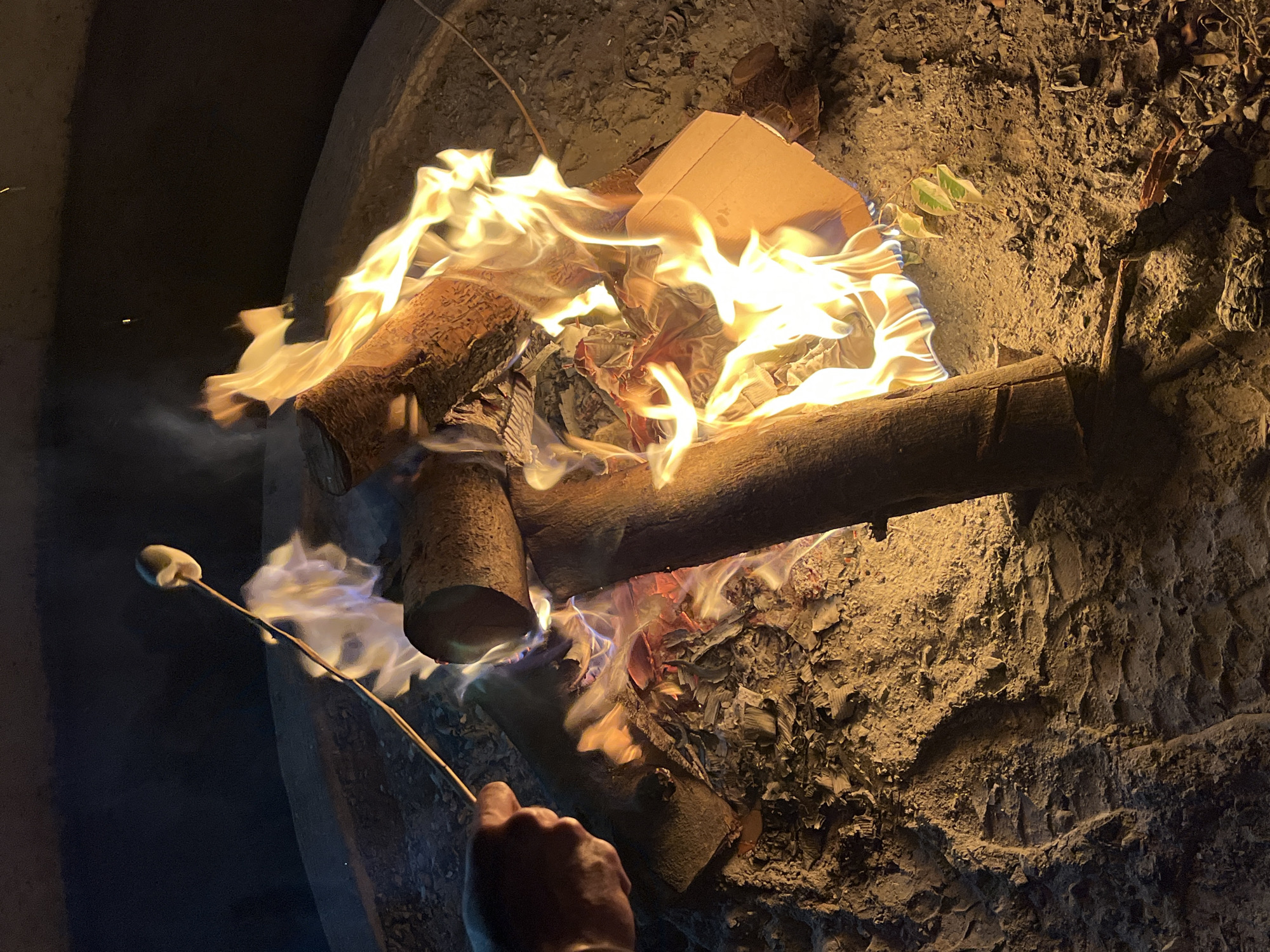 A low-angle, close-up shot depicts a campfire burning inside a fire pit. The fire pit is made of a rough, textured material that appears to be either concrete or stone. Flames engulf logs, cardboard, and paper within the pit. To the left, a hand holds a long stick with a marshmallow on the end, held over the edge of the fire. The surrounding area is dark, suggesting the fire is taking place at night. The image provides no evidence of the location being in Puebla, Mexico.