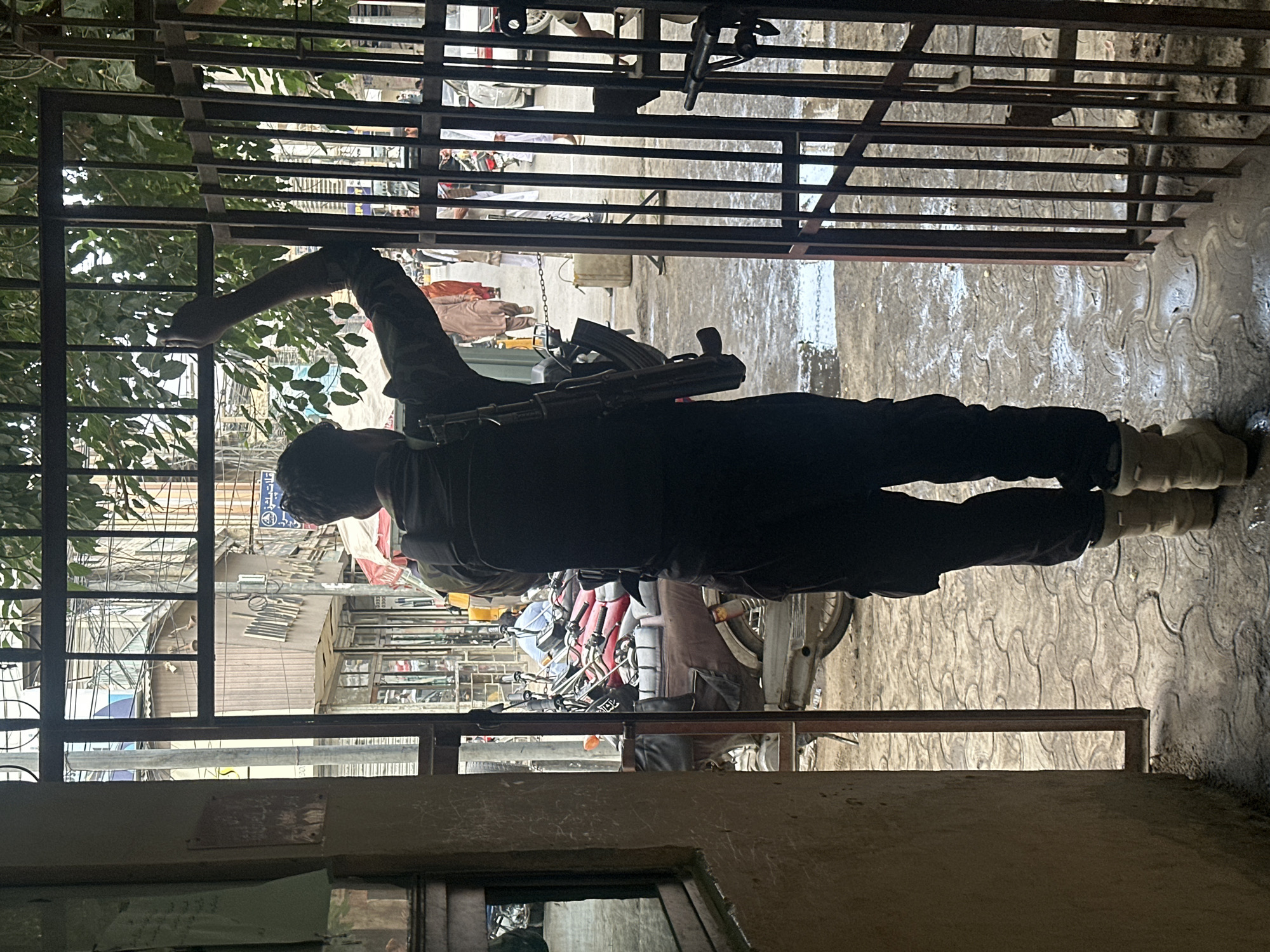 A man stands in Quetta City Tehsil, Pakistan, seen through an opening framed by a wrought-iron gate and a building structure. He carries a rifle slung over his shoulder. The street scene beyond includes motorcycles, pedestrians, and shops lining the street. The ground appears wet.