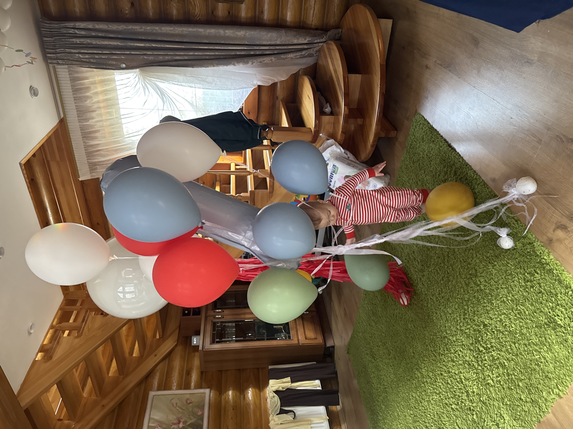 A baby or young child, dressed in a red and white striped outfit, lies on their back on a vibrant green shag rug, surrounded by a festive array of balloons and decorations. The child appears to be looking upwards, with a white bag (partially showing "JUNIORS" text, possibly diapers) positioned near their left arm.
Numerous balloons in various colors—white, light blue, red, light green, and yellow—are scattered around the child and rise towards the ceiling. Some are clustered together, tied with white ribbons and delicate, web-like streamers or netting. Two spherical white decorations, adorned with white netting and ribbons, are also visible on the rug, one close to the child's head.
The setting is an indoor space characterized by extensive use of wood, giving it a rustic or cabin-like feel. Wooden log walls, a wooden staircase with railings, and wooden furniture (including a tiered table unit and a cabinet with glass doors) are prominent. Bright natural light illuminates the room through a large window covered by sheer white and gray drapes, suggesting it is daytime. On the wooden cabinet, a digital clock displays "6:27". A framed floral painting hangs on a wooden wall.
The scene depicts a celebration, likely a birthday party or special event for the child, in a cozy home in Tomilino, Russia.