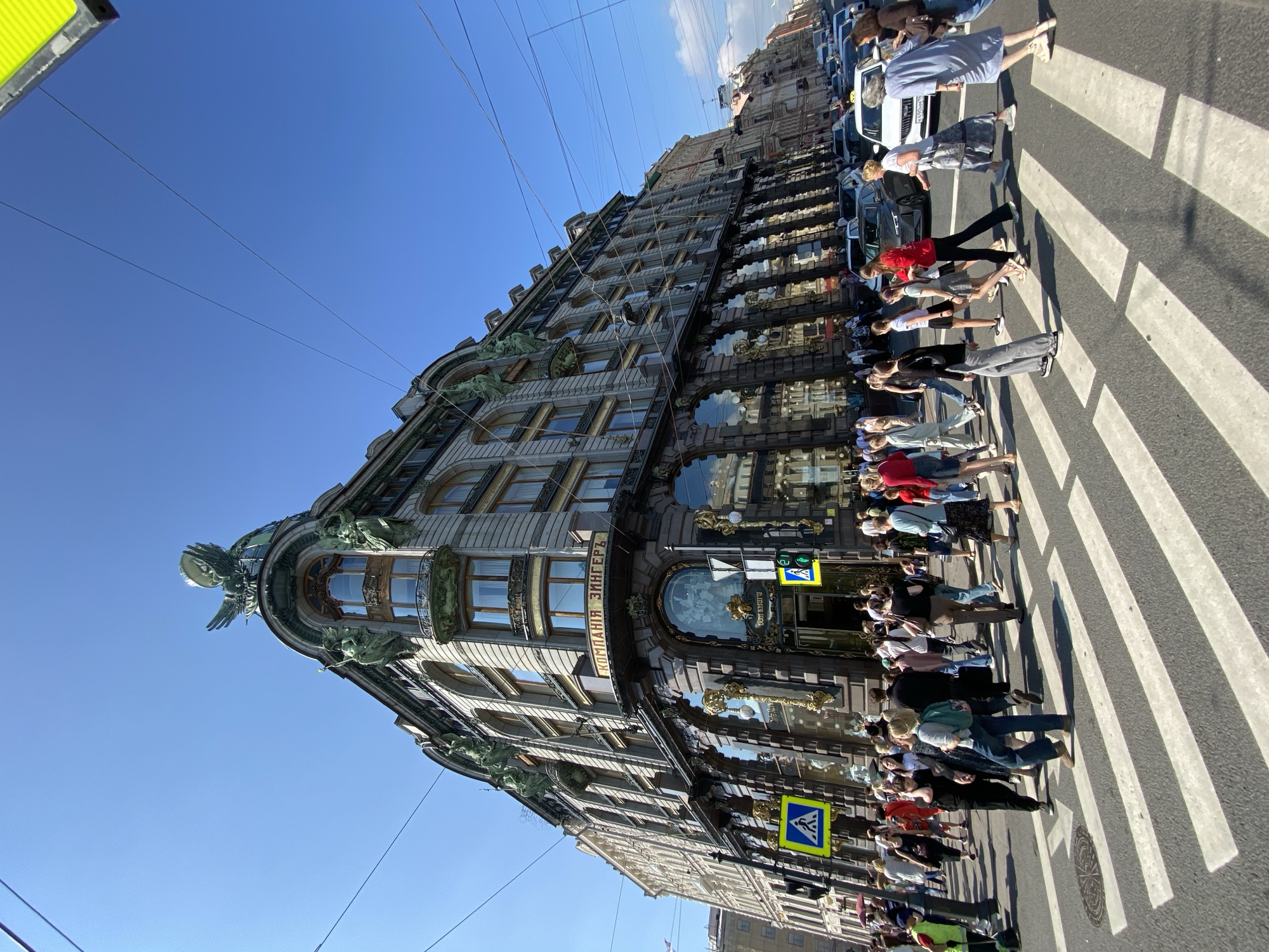 This media file depicts a group of people crossing a street at a crosswalk in front of a grand, ornate building. The building's architecture is detailed, with classical elements and statues adorning its facade. The scene is bathed in bright sunlight, suggesting a clear, sunny day, and the sky is a vibrant blue. Cars are visible on the street, indicating an urban setting.
The people are diverse in age and attire, dressed in casual summer clothing. They appear to be tourists or pedestrians going about their day. The activity is a simple crosswalk crossing. The time of day seems to be midday, given the strong shadows cast by the people and the building.
Notable details include the striking architecture of the building, which has a unique triangular shape at its corner. The presence of traffic lights and pedestrian crossing signs further contextualizes the scene as an active urban intersection. The overall mood is one of everyday life unfolding in a historic and visually impressive city.
Visible text on the building appears to be in Cyrillic script. Specifically, on the building's facade, the text "КОМПАНИЯ ЗИНГЕРЪ" is clearly legible, which translates to "Singer Company". There are also yellow square signs with a blue pedestrian symbol, indicating a crosswalk.
