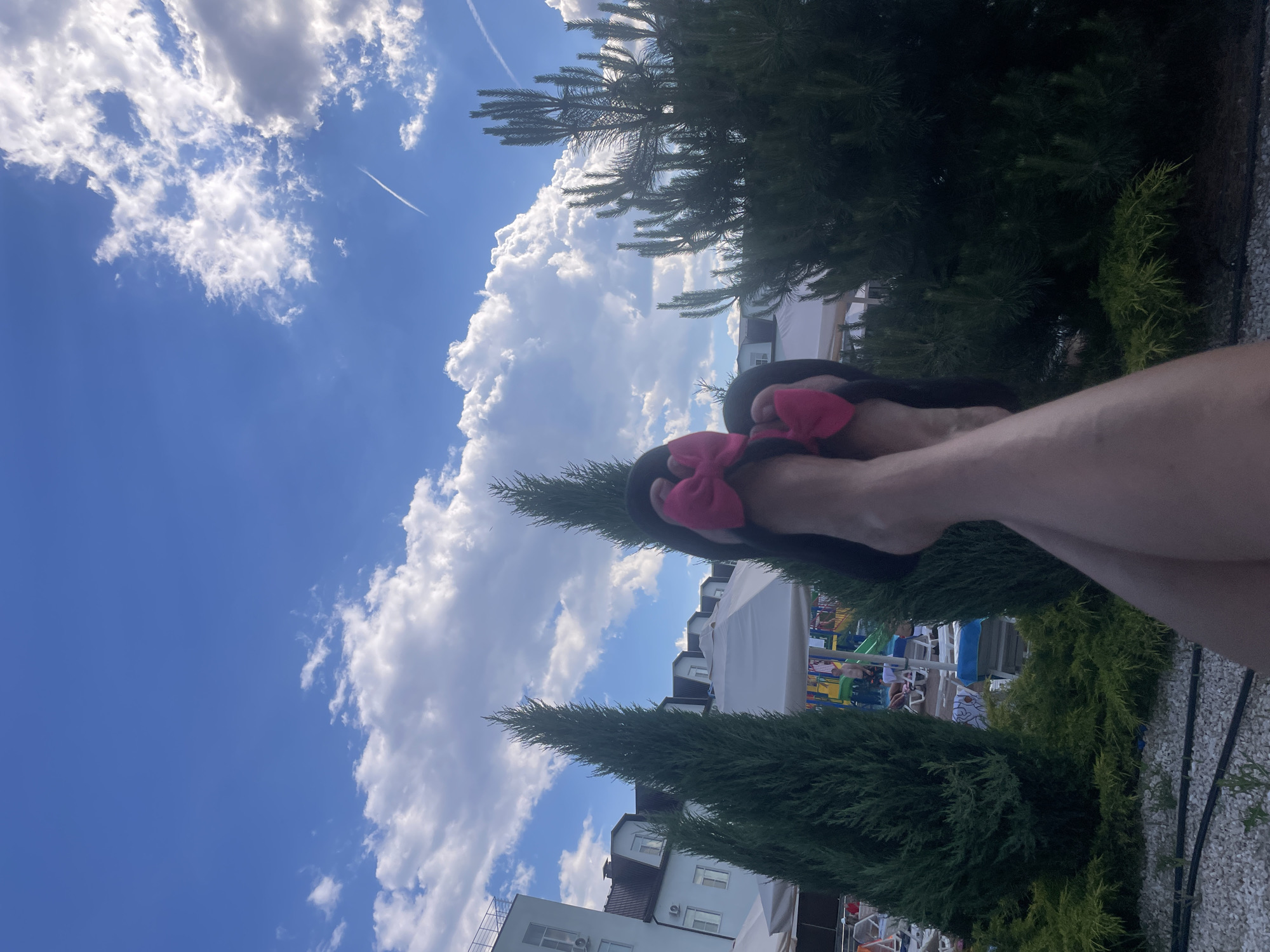 The image shows a person's legs and feet wearing black flip-flops with pink bows, crossed in the foreground. The feet are positioned against a backdrop of a blue sky with scattered white clouds and a contrail from an aircraft. In the mid-ground, there are green coniferous trees and a white building with multiple windows. Partially visible behind the trees are white umbrellas and what appears to be a playground or water park area with colorful structures and people in the distance. The ground in the foreground is covered with small white pebbles and some dark hoses or wires. The scene is outdoors, likely at a resort or recreational area in Пристанное, Russia.