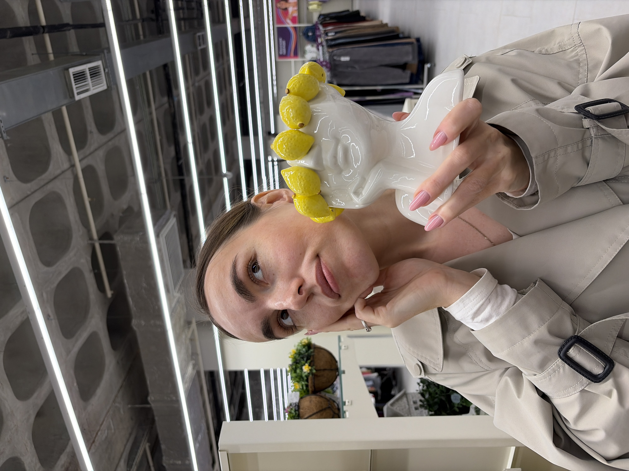 A portrait-style image, taken indoors in Izhevsk, Russia, features a young woman looking directly at the camera with a calm, direct expression. She is wearing a light beige trench coat over a white long-sleeved top. Her hair is pulled back, and her left hand, adorned with a ring, rests gently beneath her chin. Her right hand holds a distinctive white ceramic decorative object, shaped like a stylized head or bust, which is strikingly embellished with multiple bright yellow lemon-shaped ornaments along its side. Her fingernails are painted with a light pink polish.
The setting appears to be a modern commercial interior, possibly a store or showroom. The background features a high ceiling with exposed concrete structures, likely a waffle slab design, and prominent parallel linear light fixtures providing artificial illumination. Industrial-style air ducts or vents are also visible overhead. In the mid-ground, a white shelf displays two hanging planters filled with green foliage and small yellow flowers. Further back, shelves stocked with various items, possibly fabrics or other merchandise, line the walls. No discernible text is visible within the image.