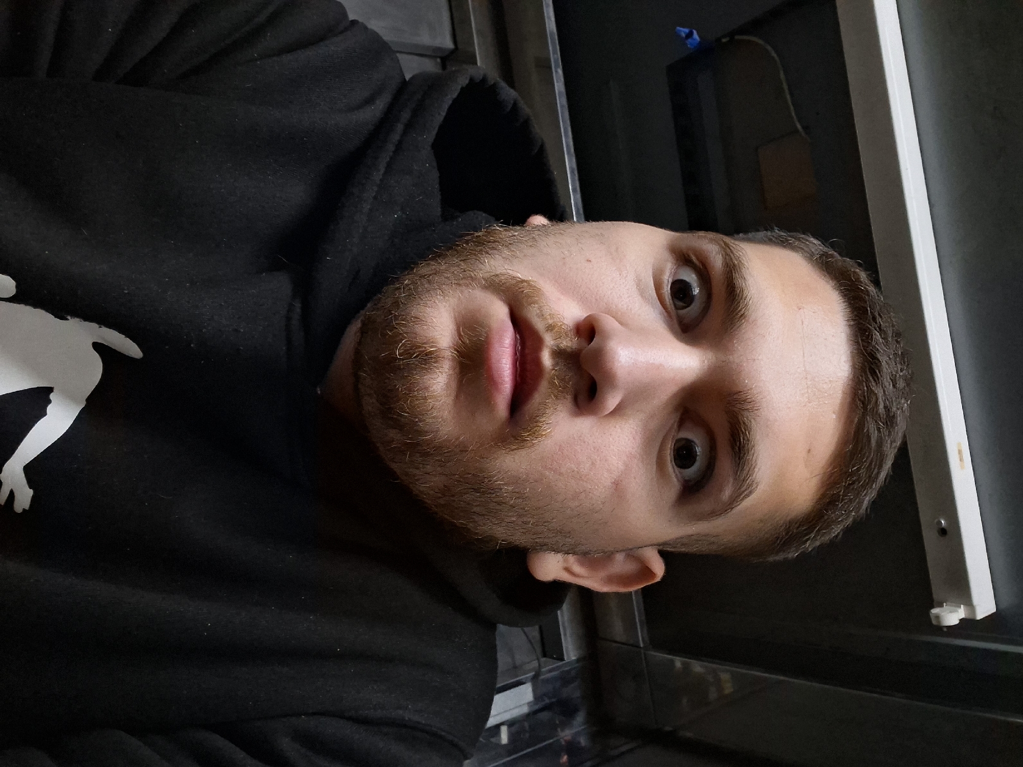 A young man with short brown hair and a light brown beard is taking a close-up, downward-facing selfie. He is wearing a black hooded sweatshirt with a white graphic on the chest. His eyes are wide and looking directly at the camera. The background appears to be indoors, with a fluorescent light fixture visible at the top and a dark, textured wall behind him. The overall impression is that of a casual, informal setting, possibly a residential or personal space. There is no clear indication of the specific location within Nicosia, Cyprus.