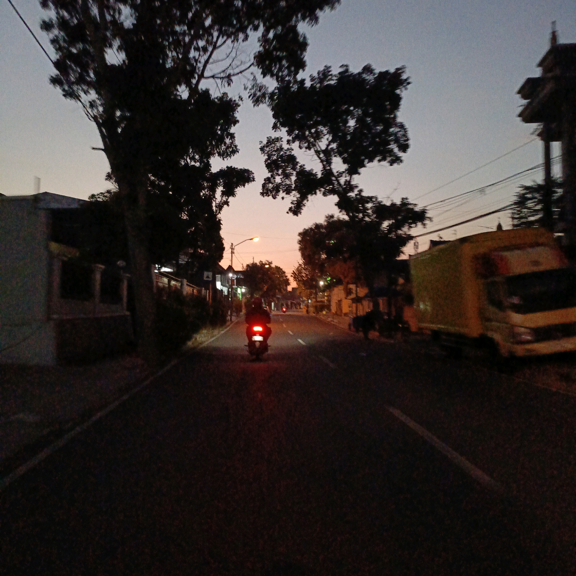 The image captures a street scene in Salatiga, Indonesia, during dusk. A motorcycle rider, illuminated by the red taillight, is traveling away from the camera down a paved road. The rider is wearing a dark helmet and jacket. On the right side of the road, a yellow truck is parked or moving slowly. The surrounding environment features silhouetted trees, buildings, and utility poles with visible wires. The sky is a gradient of pale blue and orange, indicating the twilight hours. The road surface is dark, with faint white lane markings.