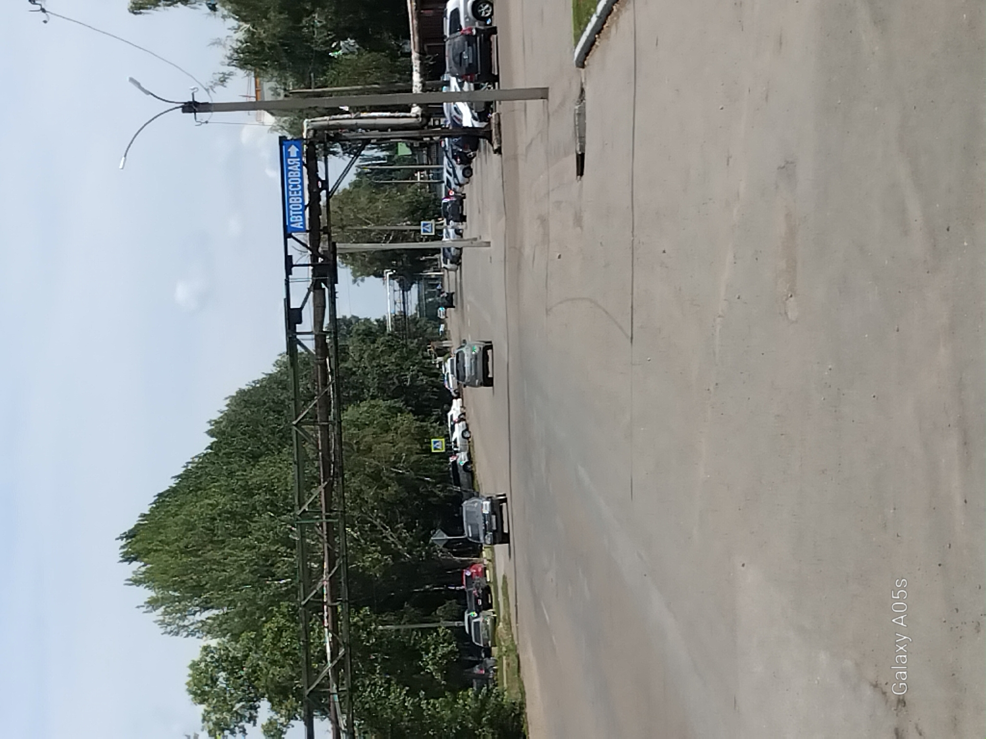 The image shows a street scene in the city of Aleysk, Russia, under a cloudy sky. In the foreground, a wide asphalt road leads into the distance, with parked cars visible on the right side. A blue sign with white lettering, reading "АВТОБЕСОВАЯ" (Autobeskova), and an arrow pointing to the right, is suspended over the road from a metal structure. A lamppost stands on the right side of the road. Trees line the left side of the street. In the background, more trees and buildings are visible. The overall scene depicts a typical urban environment on a summer day.