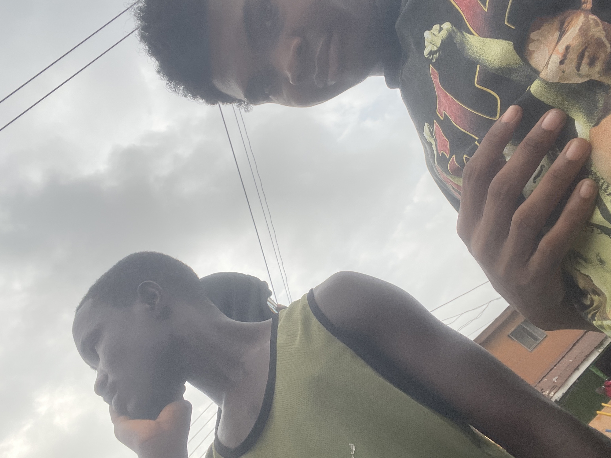 The image shows a selfie taken from a low angle, looking upwards towards a cloudy sky with power lines crisscrossing the frame. Two young Black males are partially visible. The male on the left is wearing a light green tank top with dark trim and is looking upwards with his hand near his mouth, as if he is speaking on a phone, though no phone is visible. The male on the right is closer to the camera and is looking towards the viewer, wearing a dark t-shirt with a graphic print of a person and large text. In the background, a two-story building with orange siding and a window is visible. The setting appears to be an outdoor urban environment, consistent with a location in Ado, Nigeria. The lighting is diffuse, suggesting an overcast day.