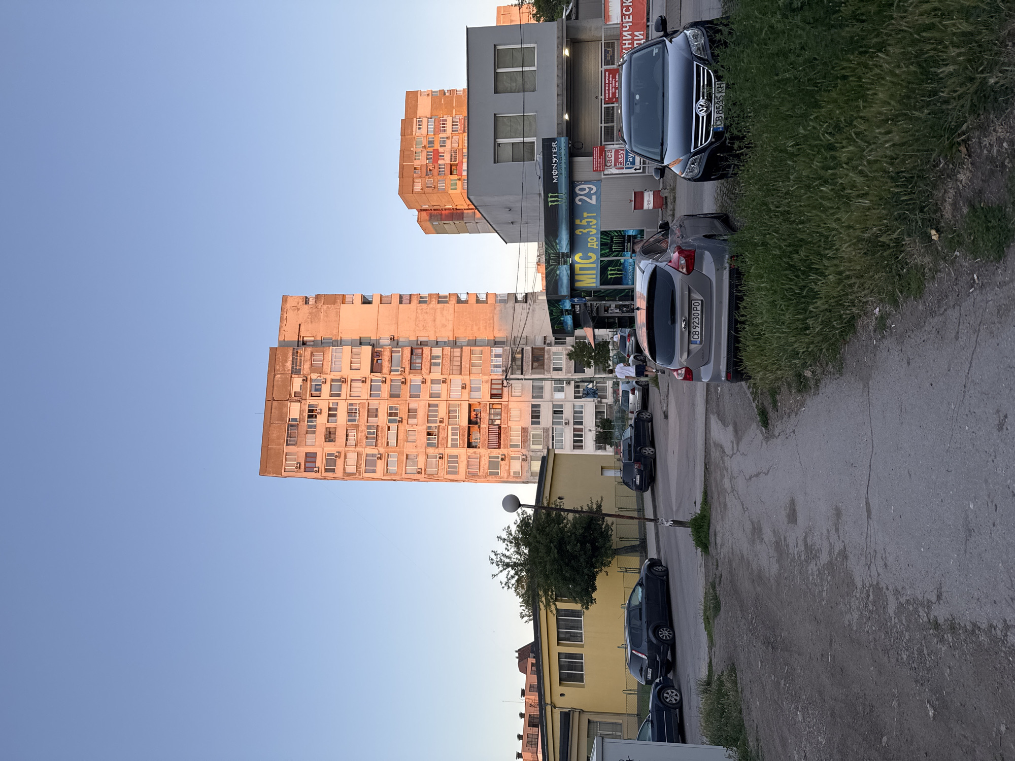 The image depicts an outdoor scene in a city, likely Sofia, Bulgaria, during what appears to be late afternoon or early evening, given the warm lighting. In the background, a tall, multi-story residential building is illuminated by the setting sun. Adjacent to it, another, shorter residential building is visible, also bathed in the warm light. In the middle ground, a commercial building with a gray facade has a prominent sign advertising "MONSTER ENERGY" and another sign in Bulgarian that reads "МПС до 3.5т 29". Several cars are parked in front of this building and on the street. A grey Ford Focus and a black Volkswagen Tiguan are prominently featured in the foreground. A street lamp stands near a tree. The ground surface consists of asphalt and dirt. A single person, a male wearing a white t-shirt and dark shorts, is walking away from the camera on the right side of the image. The sky is clear and blue.