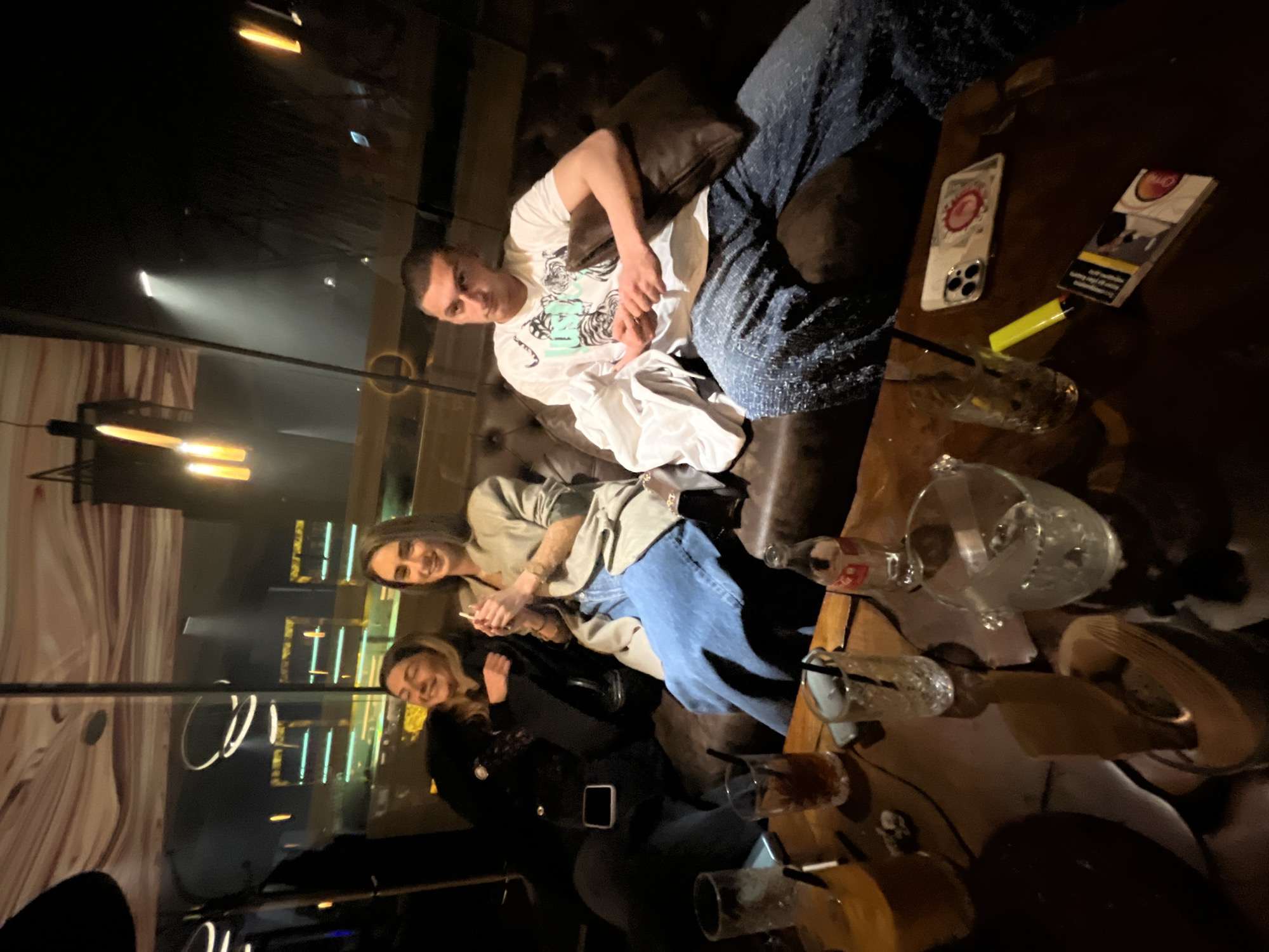 The image shows three individuals—one man and two women—seated on a dark, plush sofa within a dimly lit indoor setting, characteristic of a bar or lounge, in Burgas, Bulgaria. All three subjects are smiling and looking towards the camera, indicating a social and relaxed atmosphere.
The man, positioned on the left side of the sofa, is wearing a white t-shirt adorned with a graphic design featuring stylized tigers or cats, and blue patterned pants. Next to him, a woman with dark hair wears a light-colored jacket over a top and blue jeans, holding a smartphone. The third person, a woman with lighter hair, is seated further right, dressed in a dark top, possibly a black jacket over a lace garment, and dark pants.
A dark wooden table in front of the sofa is cluttered with various items: multiple empty or partially consumed drinking glasses with straws, some containing ice, an empty Coca-Cola bottle, and an ice bucket. Also on the table are a yellow lighter, a pack of cigarettes, and a smartphone in a clear case with a red circular design on its back. A small decorative skull is visible near the glasses.
The background is dark, featuring numerous artificial light sources and reflections, suggesting glass or mirrored surfaces. Linear and circular light fixtures are reflected, and a spotlight creates a prominent beam in the distance. The scene depicts an evening gathering.
Visible text includes the brand "ONE" on the cigarette pack, along with health warnings such as "SMOKING KILLS" and "FUMAR MATA." The man's t-shirt clearly displays a black and white animal graphic.