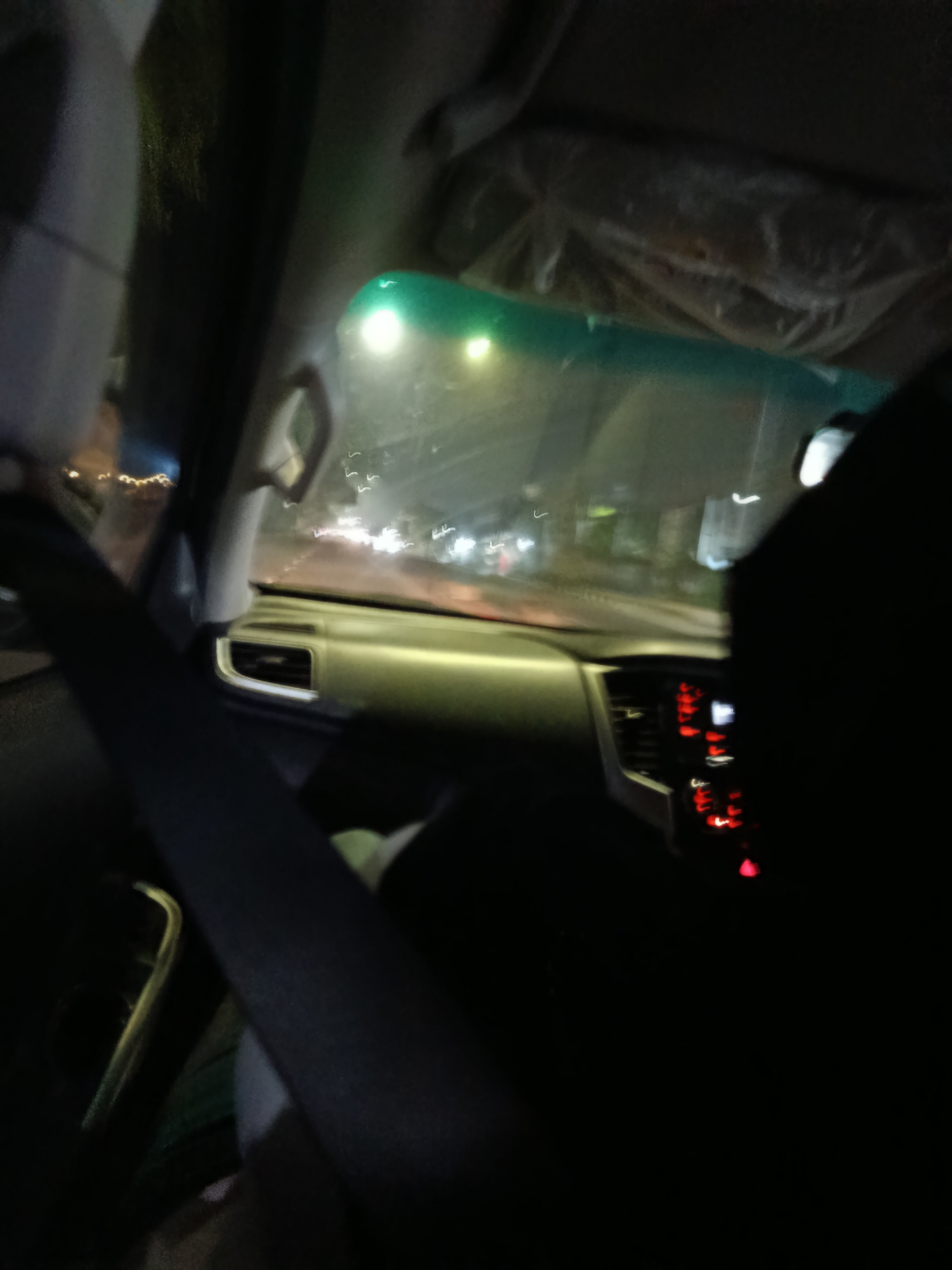 The image depicts the interior of a vehicle at night, possibly in Cibinong, Indonesia. The perspective is from the passenger seat, looking forward. The dashboard is visible, with illuminated red dials and a green tinted windshield. Streaks of light from passing vehicles are visible through the windshield, suggesting movement. A person's shoulder and arm, wearing a dark garment, are in the foreground, obscuring part of the view. The overall lighting is dim, with ambient light from the road and the vehicle's interior. The scene appears to be a snapshot of travel at night, possibly on a highway or main road.