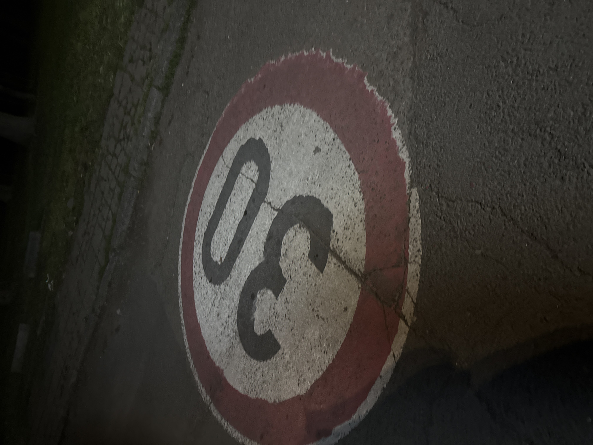 The media file presents an overhead view of a circular speed limit sign painted directly onto a dark asphalt surface. The sign features a prominent red outer ring surrounding a white interior, within which the number "30" is painted in dark black digits. This marking indicates a speed limit of 30, likely kilometers per hour, given the location context of Burgas, Bulgaria.
The asphalt road surface appears worn, with visible cracks propagating through both the painted sign and the surrounding pavement. The paint on the sign, particularly the red border, shows signs of degradation and uneven edges, suggesting age and exposure to weather. To the left side of the frame, a strip of dark green grass is visible, bordering what appears to be a cobblestone or paved path.
The lighting in the image is dim, characteristic of nighttime or very low light conditions, possibly illuminated by artificial sources like streetlights or a camera flash. The overall tone is dark and muted, emphasizing the texture of the asphalt and the painted sign. No human activity or specific weather conditions are discernible, presenting a static, functional depiction of urban infrastructure in a nocturnal setting.