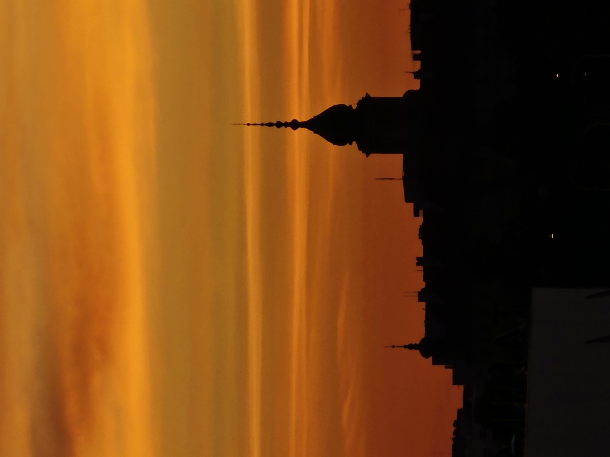 A silhouette of a city skyline, likely Warsaw, Poland, is visible against a vibrant orange and yellow sunset. The sky is filled with horizontal bands of clouds, creating a dramatic backdrop. Dominating the central part of the skyline is a distinct building with a domed roof and a tall, spire-like pinnacle. To the left of this main structure, a smaller, similarly shaped dome with a spire is also visible. The rest of the skyline is a dark, irregular mass of buildings, indicating a dense urban environment. The lower portion of the image shows a lighter, flat structure in silhouette, possibly a rooftop or another building in the foreground. The overall impression is of a cityscape at dusk, with the warm colors of the sunset casting a rich glow.