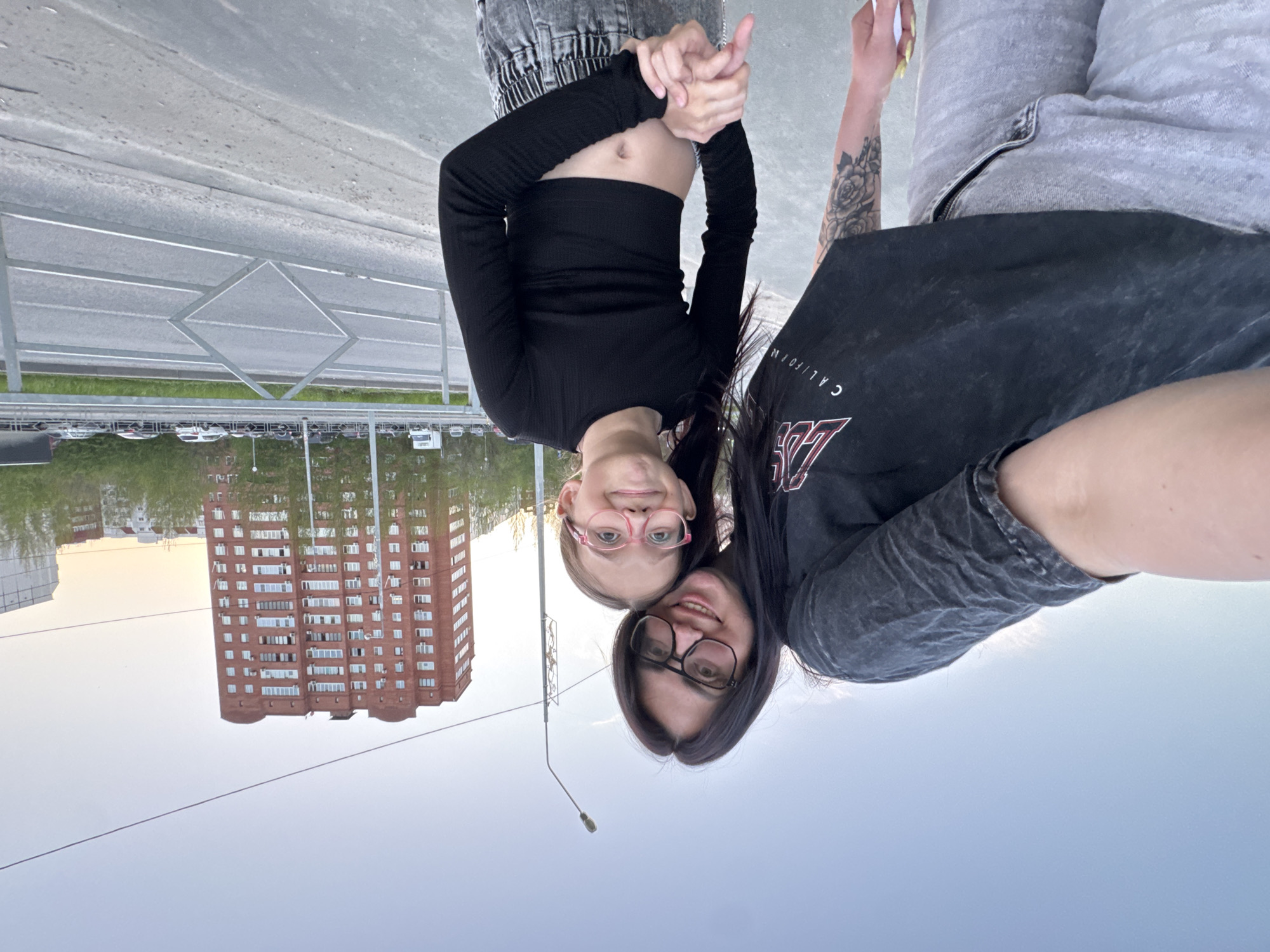 Two people are taking a selfie outdoors. One individual is wearing a black long-sleeved crop top and light gray jeans. The second person has a tattoo on their arm and is wearing a gray T-shirt and gray jeans. Both are wearing eyeglasses. In the background, there is a tall brick building and a gray road with a metal railing. The sky is light blue.