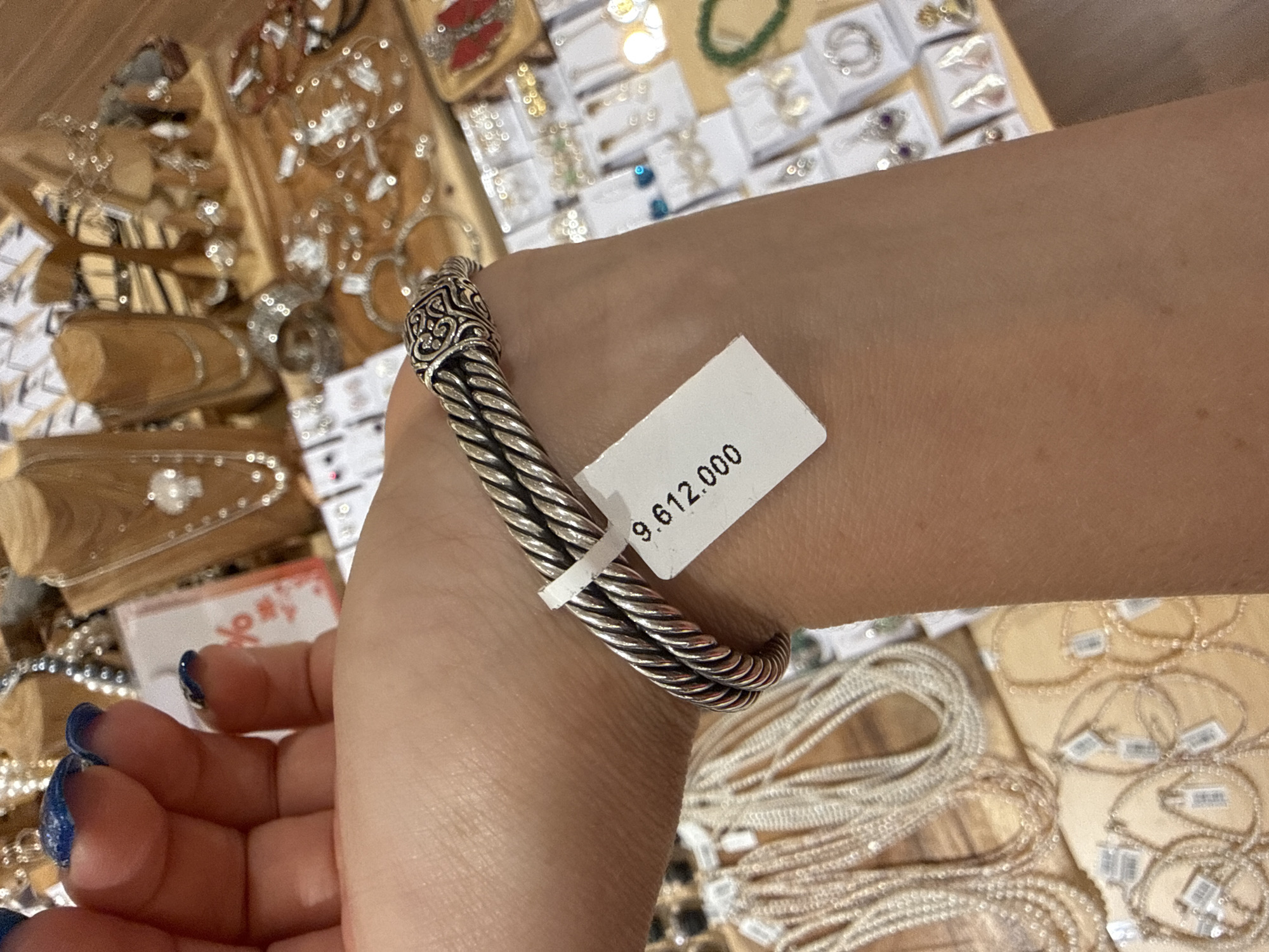 The image shows a close-up of a person's left wrist and forearm, adorned with a silver cable bracelet. A white price tag with black text reading "9.612.000" is attached to the bracelet. The background is blurred but appears to be a retail display of jewelry, with various necklaces, earrings, and other accessories visible on shelves and stands. The lighting suggests an indoor environment, likely a store. The person's fingernails are painted with blue glitter polish. There is no identifiable geographical location within the image.