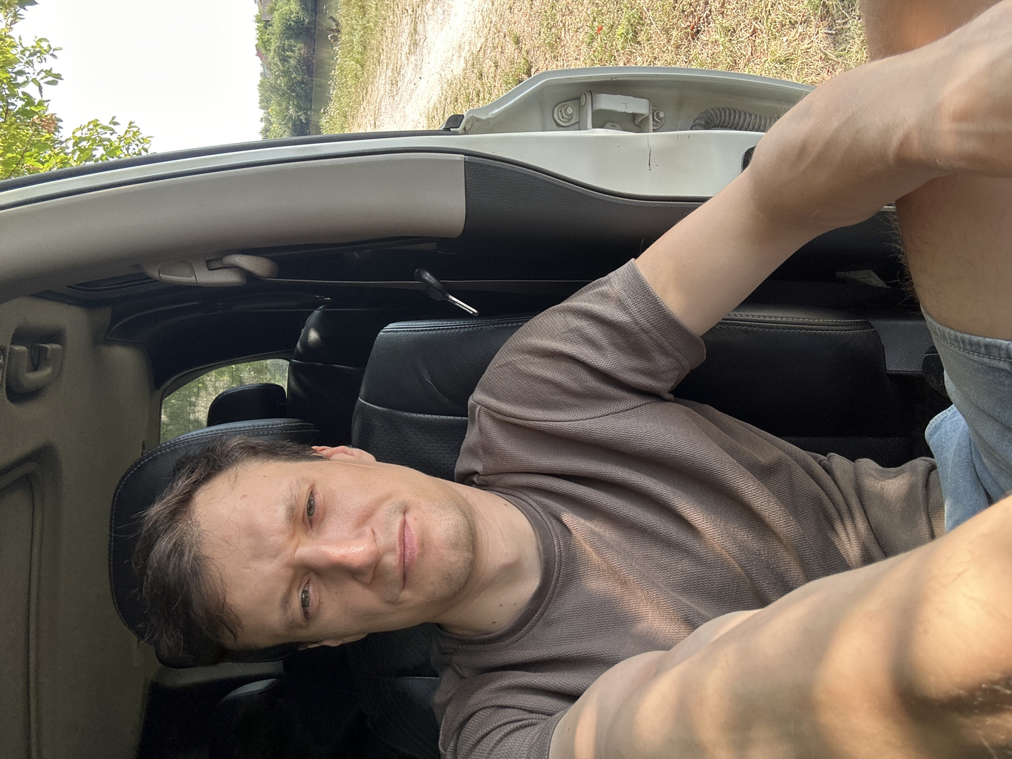 A Caucasian male is sitting in the driver's seat of a white SUV. He has short brown hair and light skin. He is wearing a brown t-shirt and blue jean shorts. The car is parked on a dirt road next to a body of water, with trees and vegetation in the background. The scene appears to be outdoors, possibly in a rural area outside of Makiv, Ukraine. The sunlight is casting shadows on the man's face and arm.