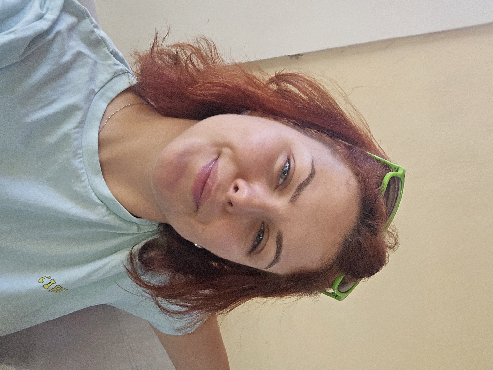 A medium shot shows a young woman with auburn hair, wearing a light blue t-shirt and green sunglasses perched on her head. She is looking at the camera and smiling. The background is a plain, light yellow wall. The image is a selfie taken by the woman.