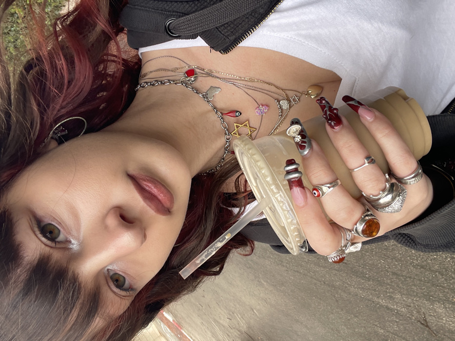 A young woman is depicted from the chest up, positioned slightly to her left, looking towards the camera. Her hair is dark brown with bangs, featuring reddish-auburn highlights in the longer sections visible to her right. She has light skin, subtle eye makeup with a shimmer on her inner eyelids, and brownish-pink lip color.
She wears a white top underneath a dark, likely black or charcoal grey, zippered hoodie or jacket. Her visible left ear features a silver-toned hoop earring with a small dark charm. She wears multiple layered necklaces, including a thicker silver chain adorned with charms such as a golden Star of David, a red teardrop-shaped hamsa, a silver map of Africa, and a small red heart. Thinner chains feature additional charms, including a pink hamsa, a small Star of David, and a golden heart-shaped locket.
Her right hand holds a clear plastic cup with a lid, containing a light brown beverage, likely iced coffee, with a clear plastic straw. Her nails are long and almond-shaped, featuring intricate nail art predominantly in dark red and silver. Specific designs include a French tip on the thumb, silver coiled elements on the index finger, a silver base with dark red abstract shapes and a small jeweled charm on the middle finger, dark red abstract designs on the ring finger, and a V-shaped silver design on the pinky finger. Numerous silver-toned rings adorn her fingers, including a ring with a red 'evil eye' on her index finger, large rings with amber-colored stones, and several plain or textured silver bands.
The background is out of focus, showing a paved surface on the left and indistinct green foliage or outdoor elements on the right, indicating an outdoor urban setting. Based on the cultural symbols present (Star of David, Hamsa), the scene is located in the city of Raanana, Israel.