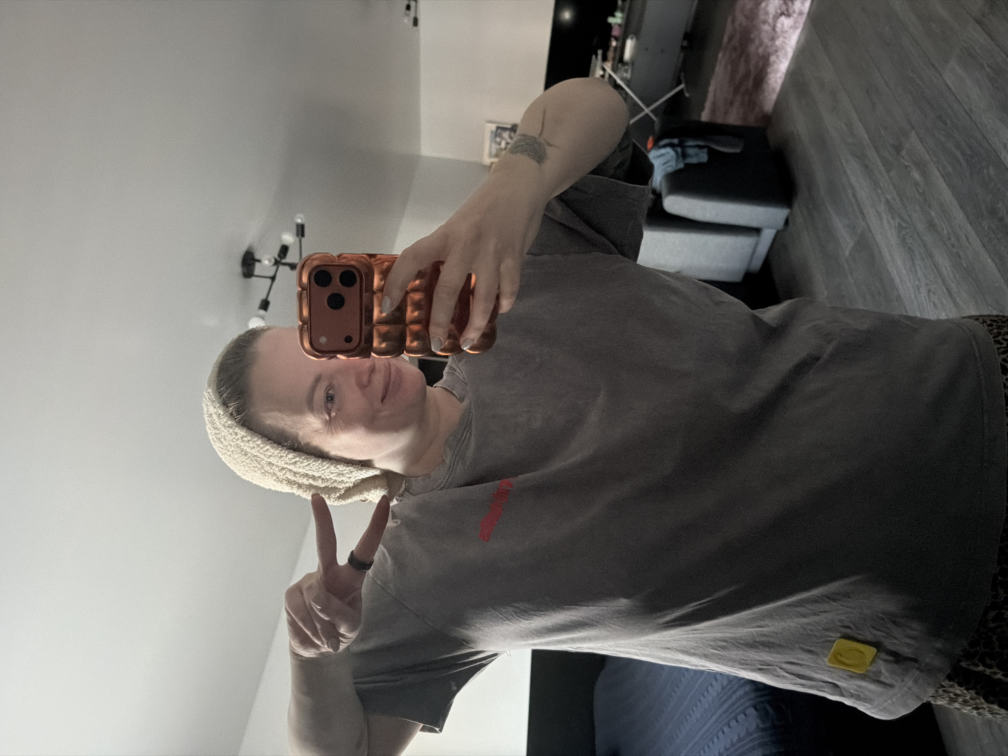 A female subject is depicted in a mirror selfie taken indoors, within a residential apartment in the city of Moscow, Russia. The subject, an adult female with light-colored hair partially covered by a cream-colored knitted head covering, is smiling and looking towards the smartphone she holds in her right hand. The smartphone, encased in a textured reddish-copper protective cover, is equipped with three camera lenses. Her left hand is raised, forming a 'peace sign' gesture. She wears a dark grey short-sleeved t-shirt featuring a horizontal red graphic on the chest and a small yellow square tag near the bottom left hem, and patterned bottoms. A tattoo is visible on her right forearm. The background, reflected in the mirror, shows a contemporary residential interior with light-colored walls, dark wood-look flooring, and a multi-bulb ceiling light fixture. Visible furniture includes a bed with dark blue bedding on the left, and on the right, a grey sectional sofa, a dark entertainment console with a television, and a light-colored rug.