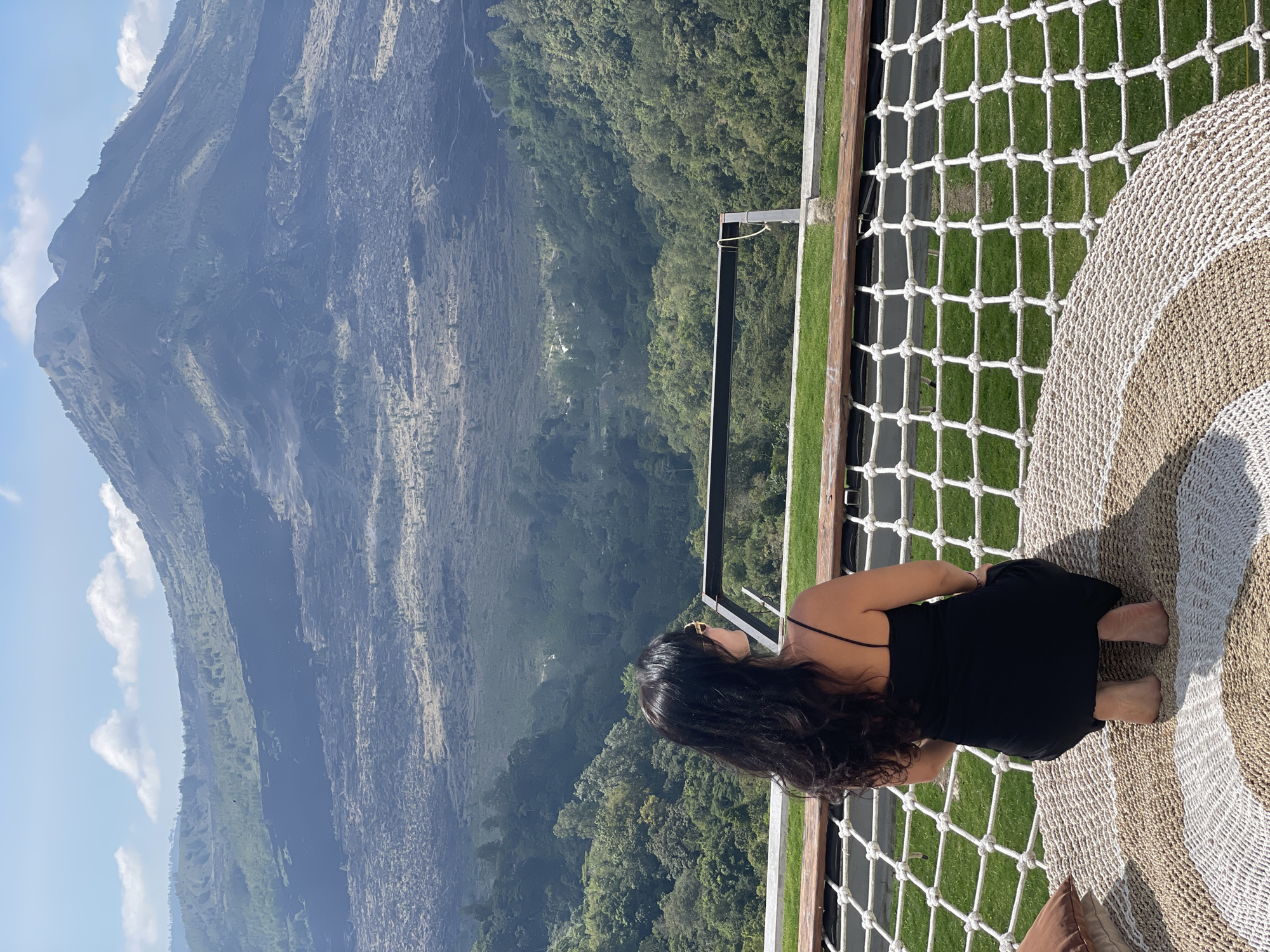A woman with long, dark hair, wearing a black slip dress and sunglasses, kneels on a circular woven mat. She faces away from the camera, looking out at a volcanic landscape in Kintamani, Indonesia. The foreground features a rope net stretching across a wooden platform, with a grassy area beyond. In the background, a large volcano dominates the scene, with a textured slope of dark volcanic rock descending into a verdant valley. The sky is blue with scattered white clouds.