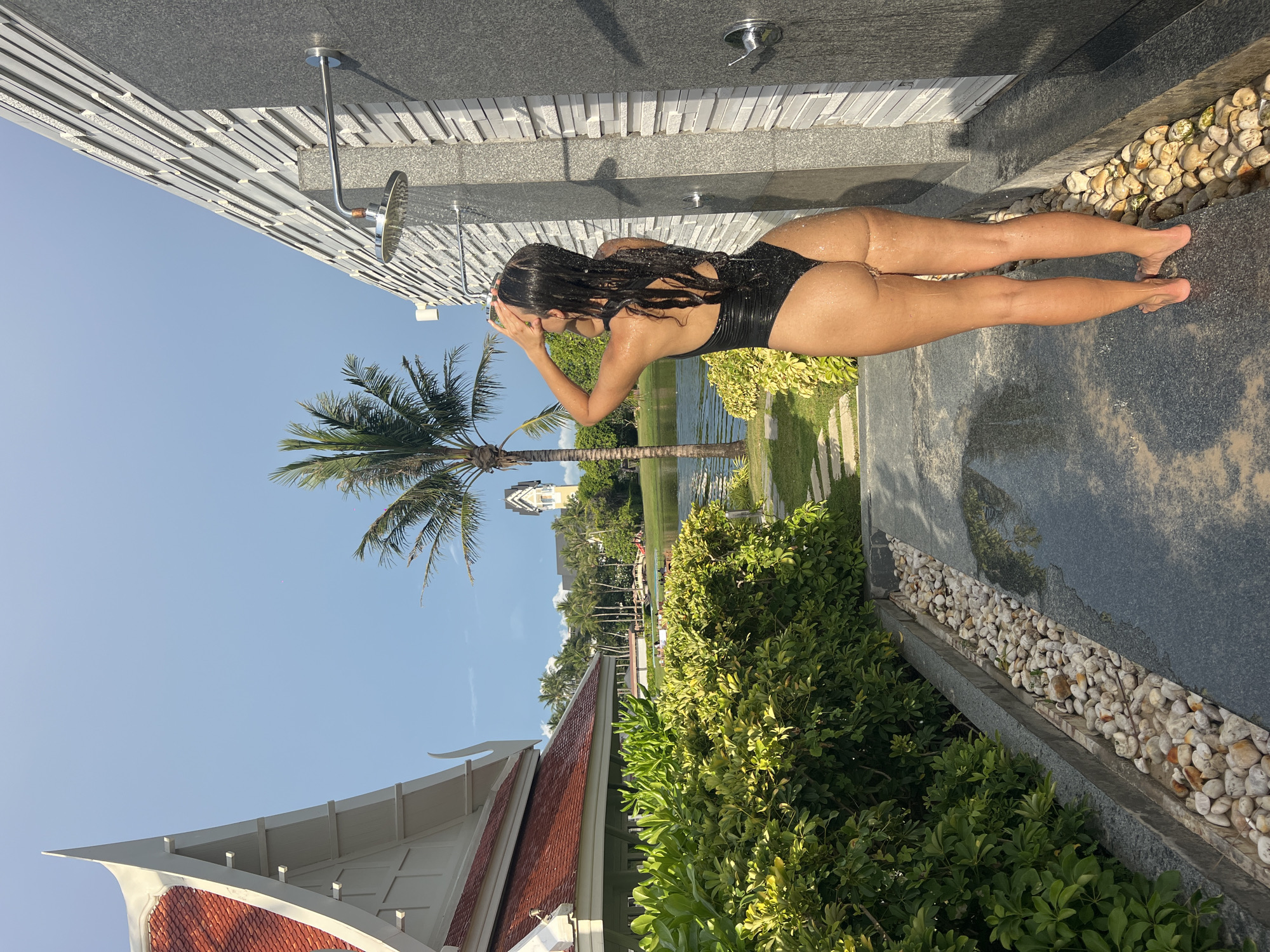 A woman in a black swimsuit stands beneath an outdoor shower in Choeng Thale, Thailand. The shower fixture is attached to a textured, gray tile wall, with a decorative border of small, smooth stones. The ground is a smooth, gray slab, wet from the shower's use. The woman is turned away from the viewer, her hands covering her face as she rinses her hair. Lush green foliage surrounds the shower area, and a palm tree stands tall in the background. A traditional Thai-style building with an ornate, red-tiled roof is visible to the lower left. A body of water and other buildings are visible further in the distance under a clear blue sky.